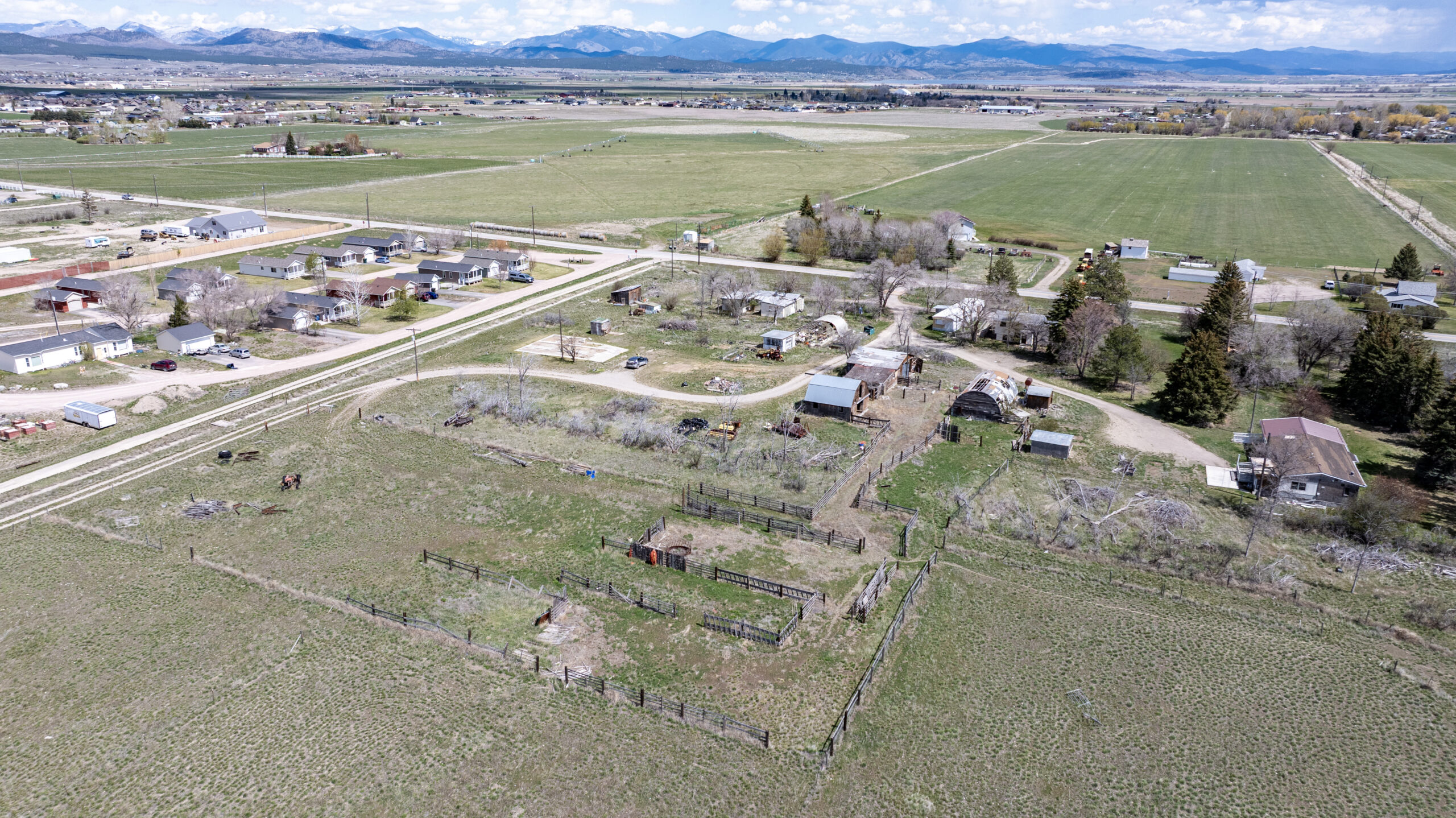 Aerial view of a rural area with fenced fields, scattered houses, barns, trees, dirt roads, and open farmland. Mountains with snow on the peaks are visible in the distance under a partly cloudy sky.