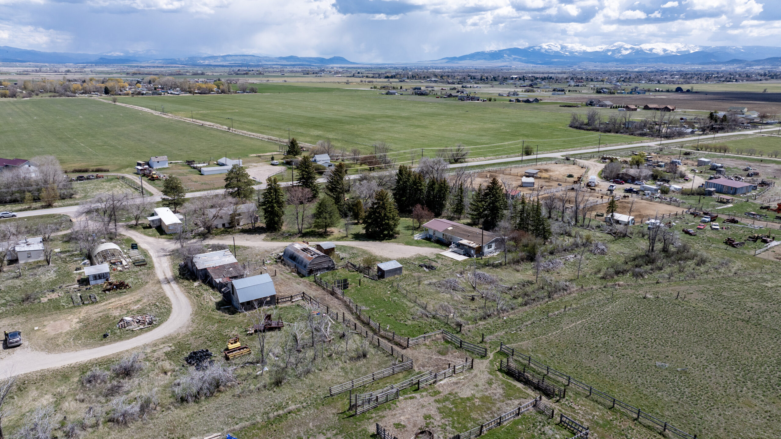 Aerial view of a rural landscape with scattered homes, barns, fenced areas, open fields, and distant mountains under a partly cloudy sky. Roads and patches of trees are visible throughout the scene.