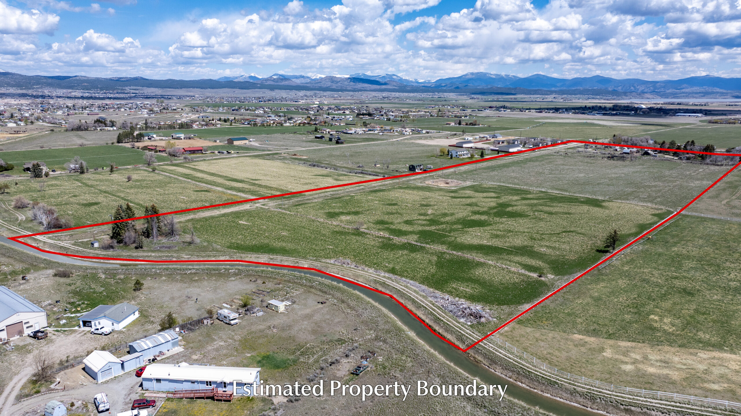 Aerial view of rural farmland with an estimated property boundary outlined in red. The land is mostly grassy fields with scattered trees, some nearby houses, and mountains visible in the distance under a partly cloudy sky.