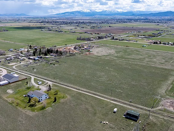 Aerial view of a rural landscape with open fields, scattered houses, roads, and a distant mountain range under a partly cloudy sky.