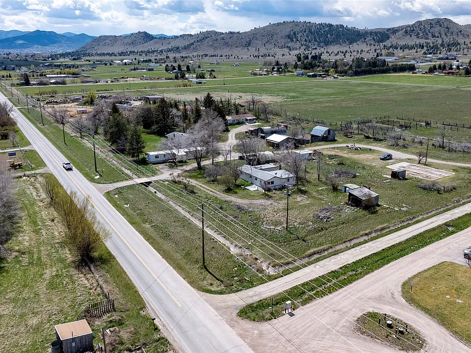 Aerial view of a rural neighborhood with scattered houses, open fields, trees, and mountains in the background. A paved road runs alongside the properties, and the landscape is green under a partly cloudy sky.
