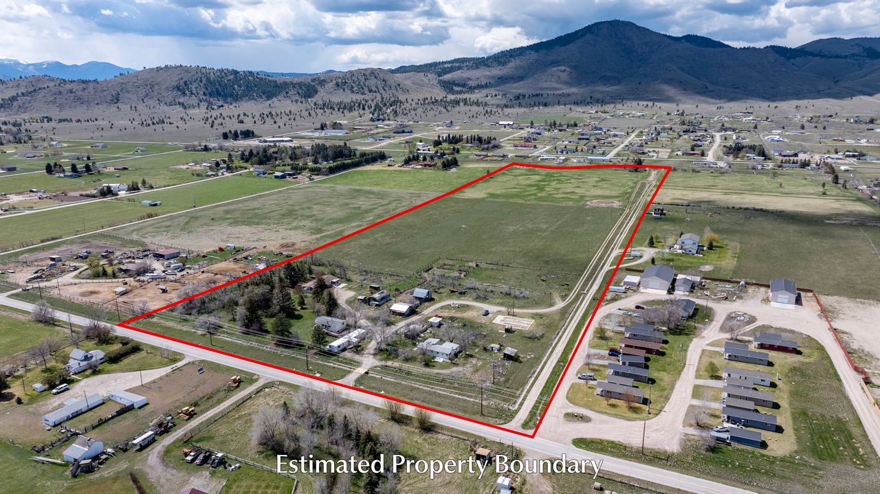 Aerial view of a large rural property outlined in red, showing open fields, scattered houses, and nearby homes, with mountains in the background under a partly cloudy sky. Text at the bottom reads Estimated Property Boundary.