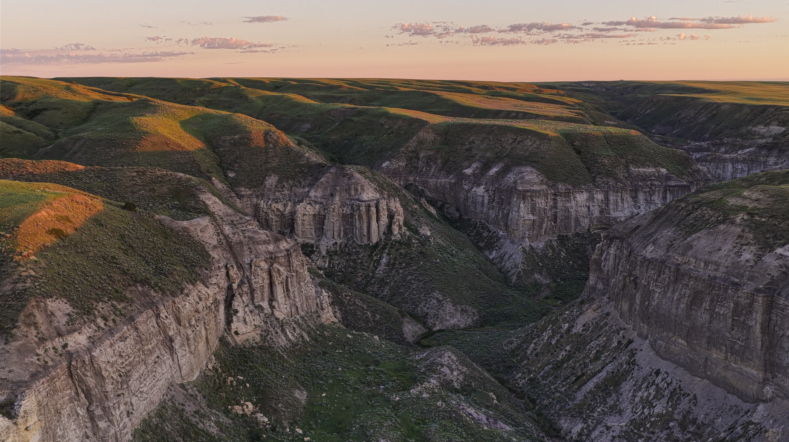 A wide canyon with steep, layered cliffs and green hills at sunset, bathed in soft, warm light under a partly cloudy sky.