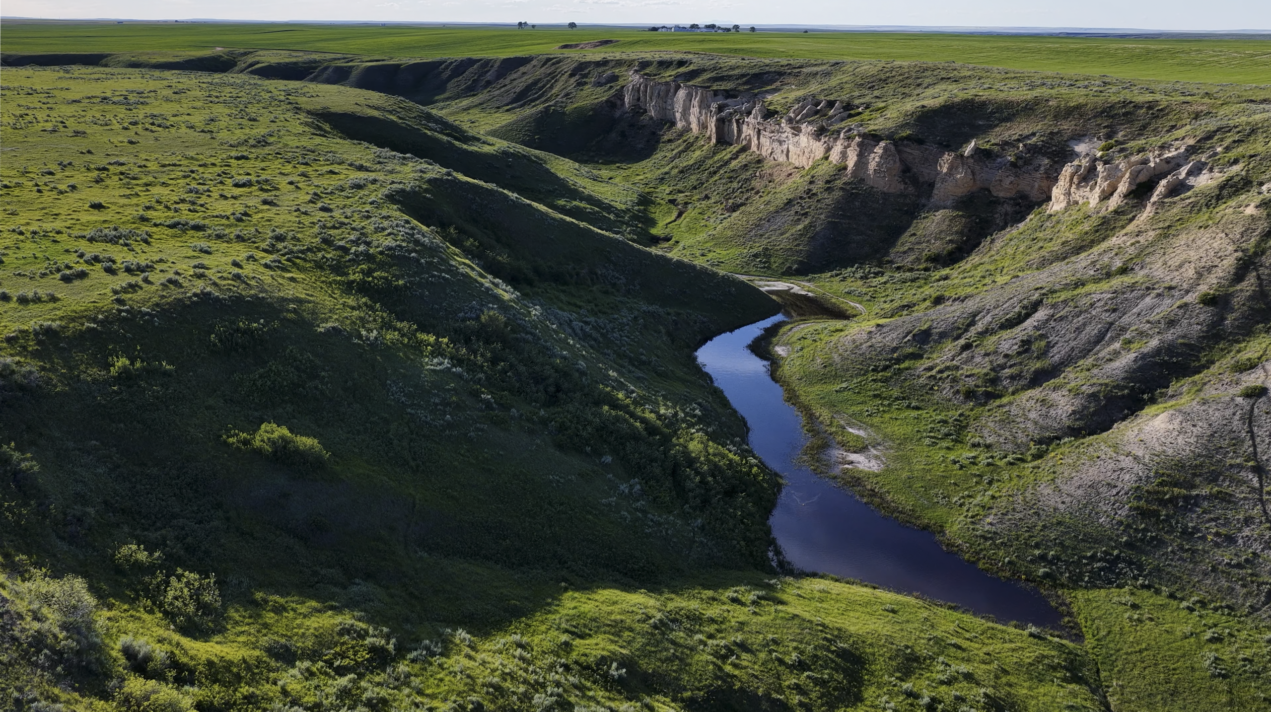 A winding river flows through a deep, green valley with steep rocky cliffs on both sides under an open sky. Grass and shrubs cover the landscape.