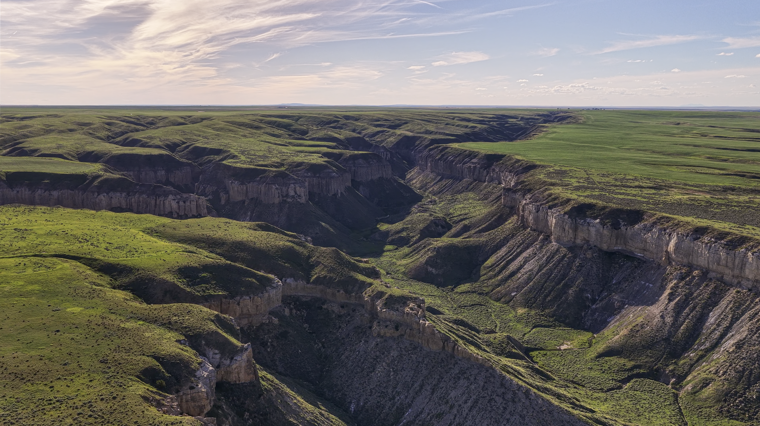 A wide canyon with steep, rocky cliffs and green grass covering the landscape under a partly cloudy sky.
