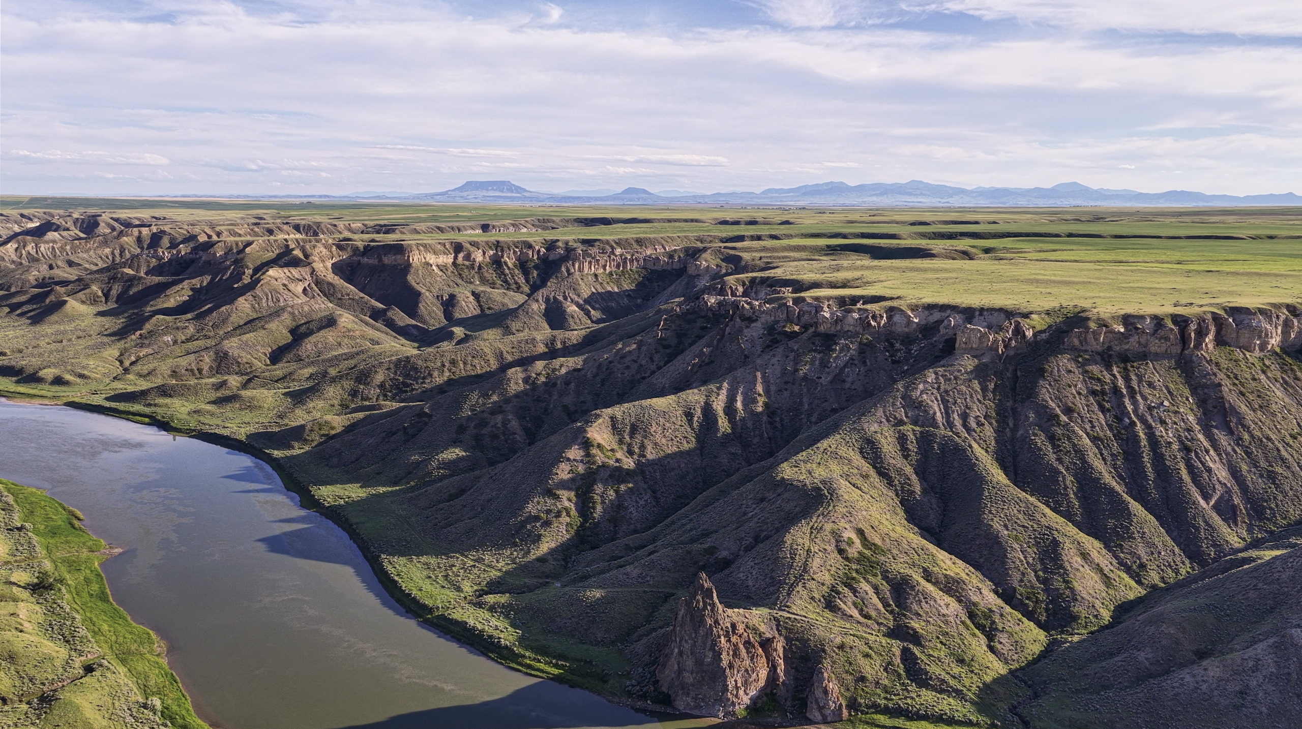 A wide river curves through a deep canyon with steep, rocky cliffs and green plateaus under a partly cloudy sky with distant mountains.
