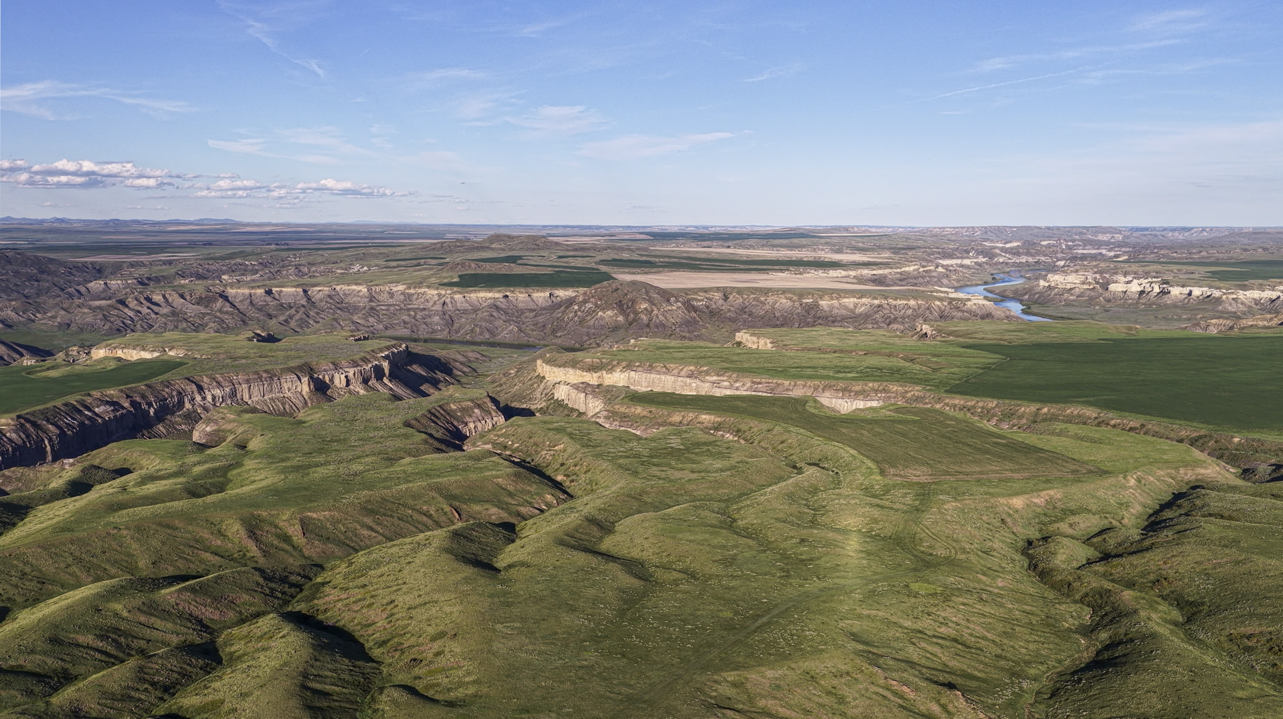 Aerial view of green rolling hills and deep canyons under a blue sky with scattered clouds, with a river winding through the landscape in the distance.