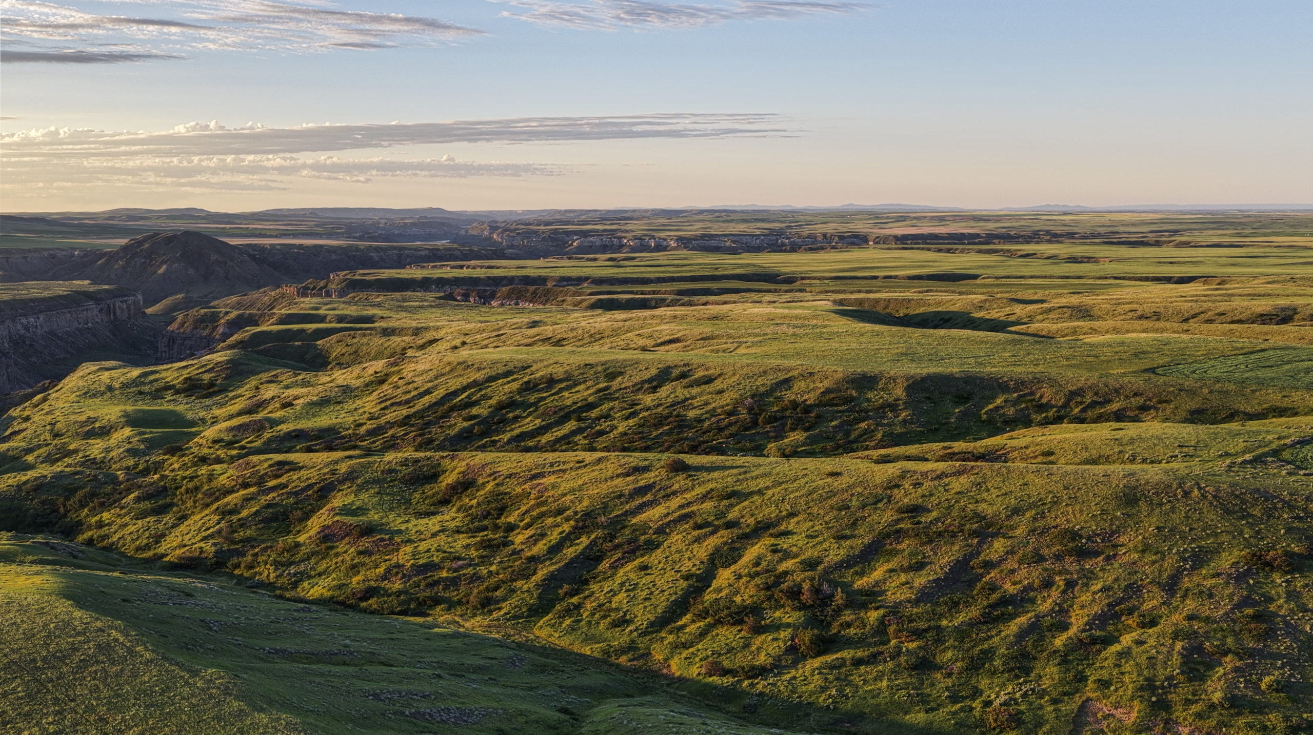 Wide landscape view of rolling green hills and valleys under a clear sky at sunset, casting long shadows across the terrain.