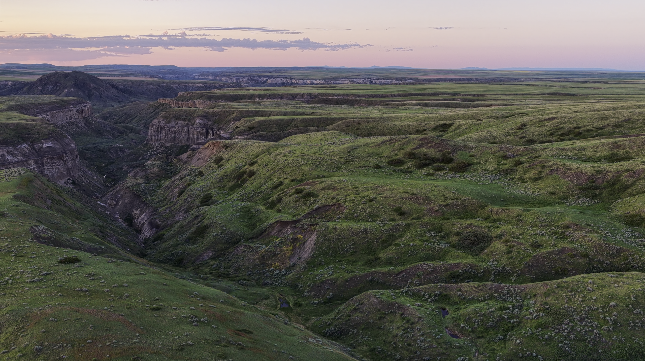 Rolling green hills and deep ravines stretch across a wide landscape under a pink and purple sunset sky, with distant plains visible on the horizon.