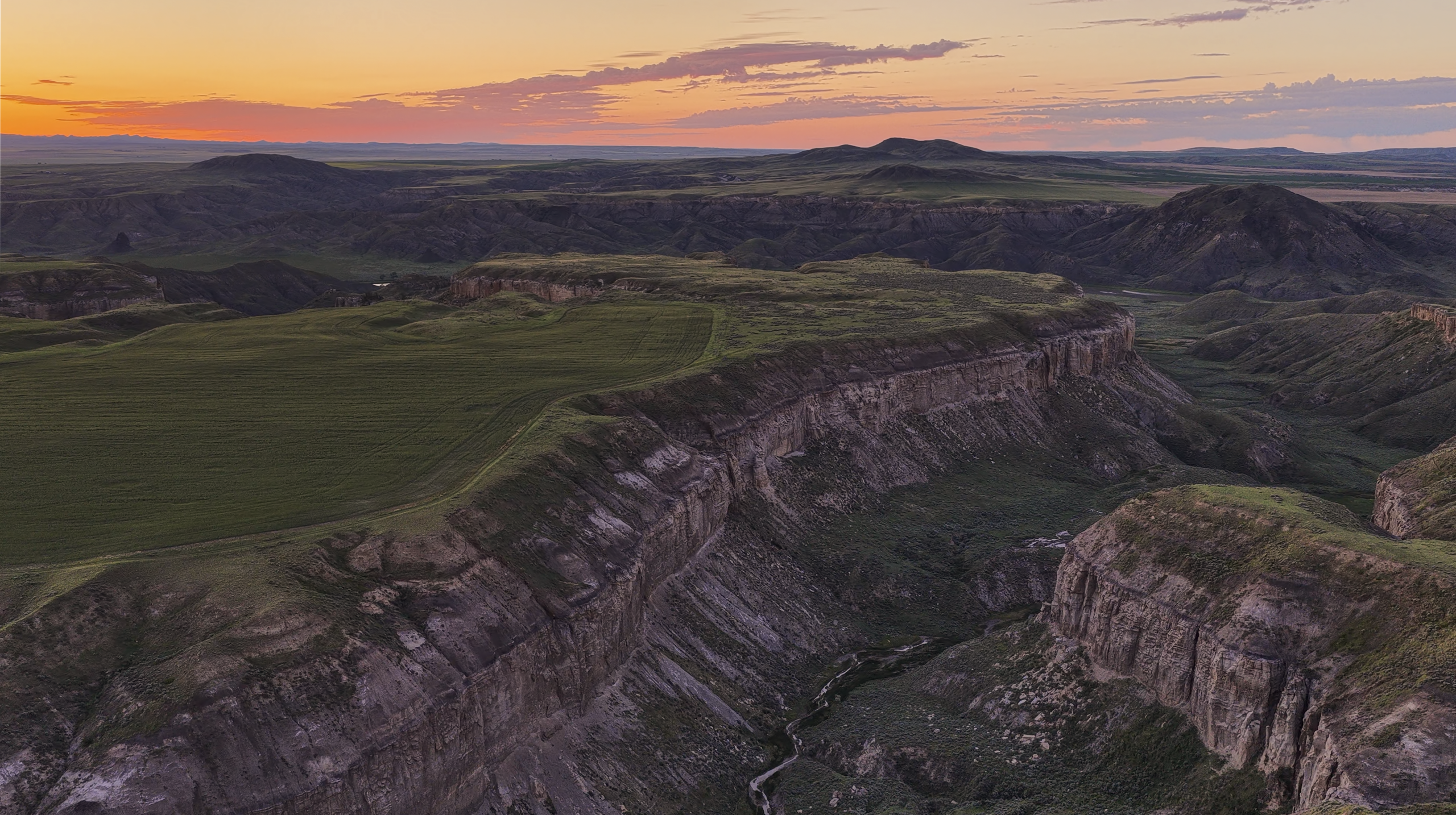 A scenic view of a deep canyon with green plateaus at sunset, featuring layered rock formations, distant hills, and a colorful sky with orange and purple hues.