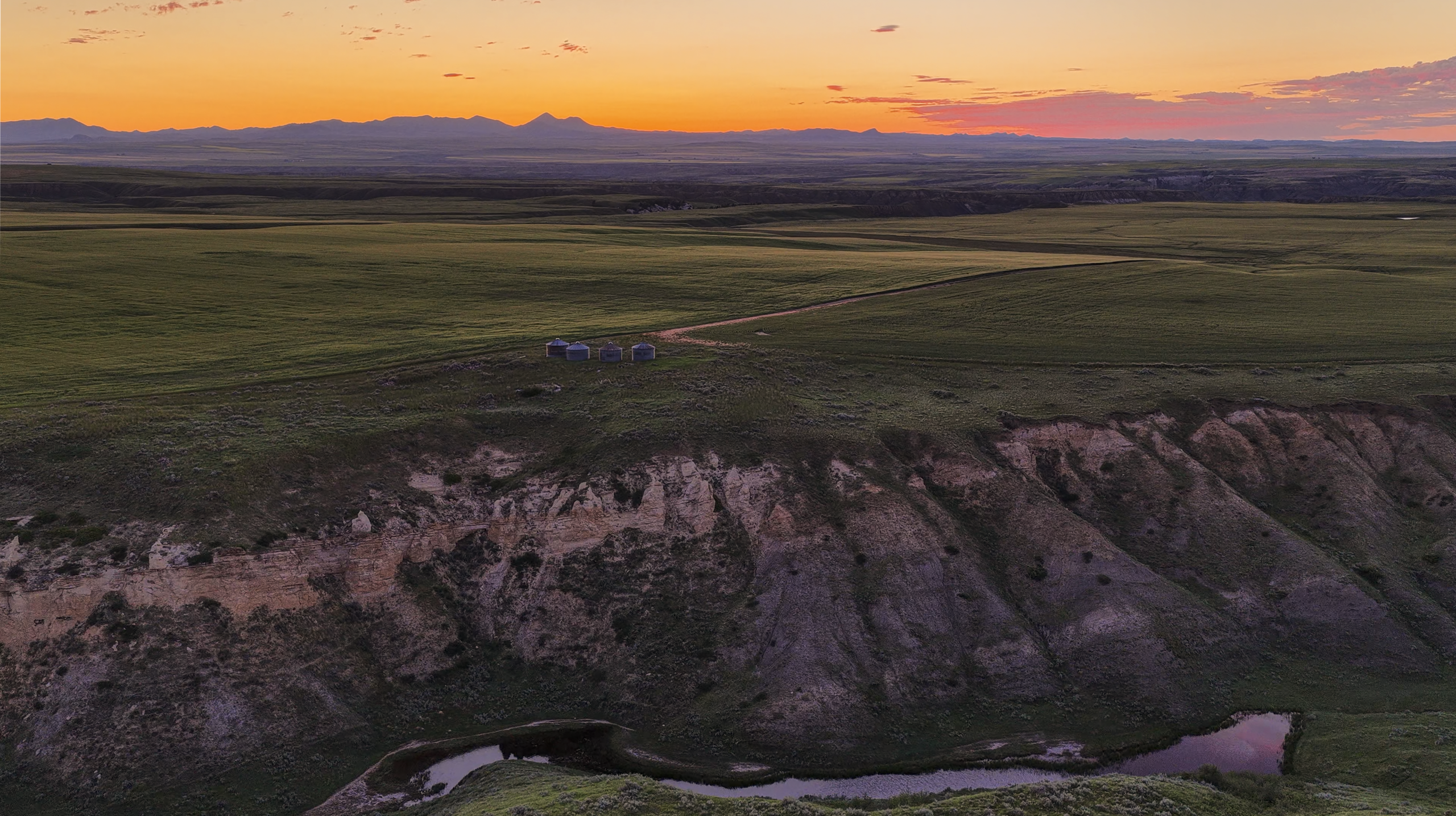 A scenic sunset over grassy plains, a rugged cliff, and a small distant building, with mountains silhouetted against an orange and purple sky.