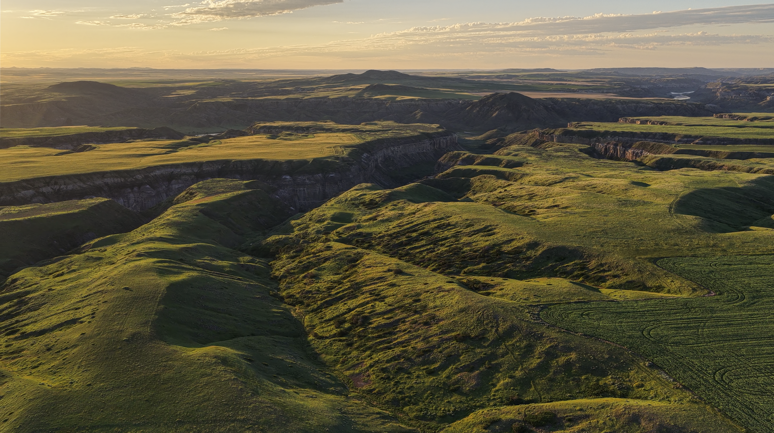 Aerial view of rolling green hills and dramatic cliffs under a partly cloudy sky at sunset, creating a scenic landscape with long shadows.