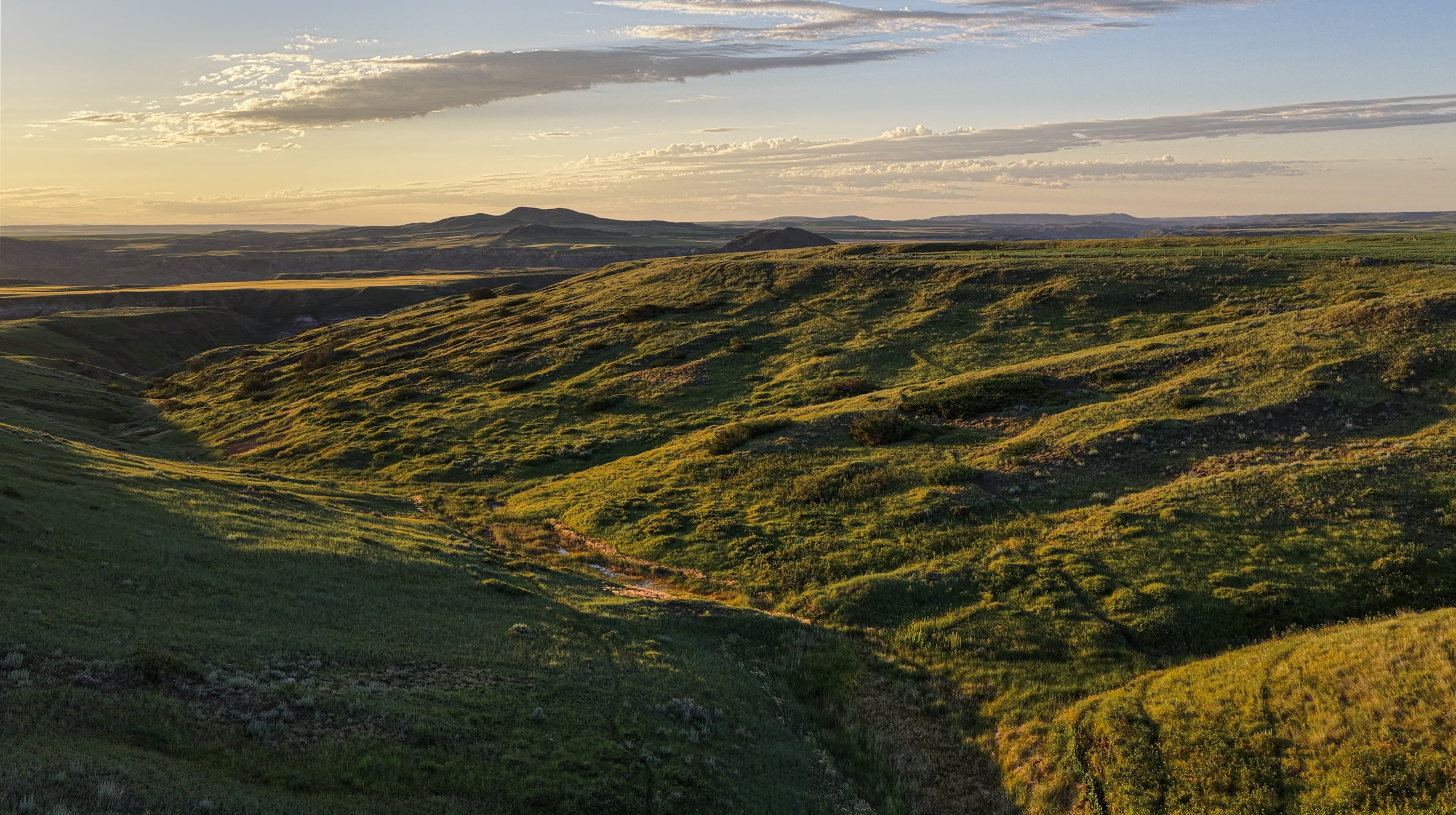 Rolling green hills are bathed in golden sunlight under a partly cloudy sky, with distant mountains visible on the horizon.