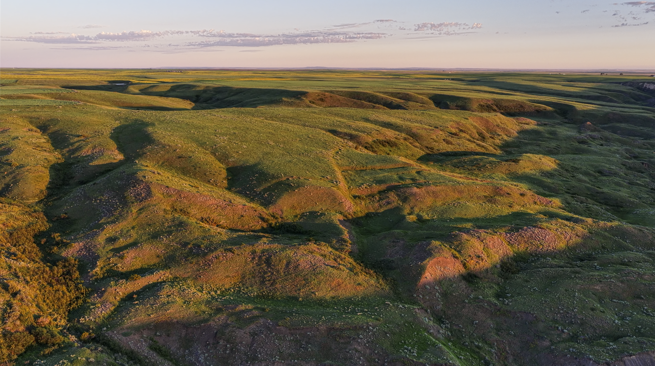 Aerial view of rolling green hills at sunset, with patches of sunlight and shadow highlighting the gentle slopes under a clear sky.