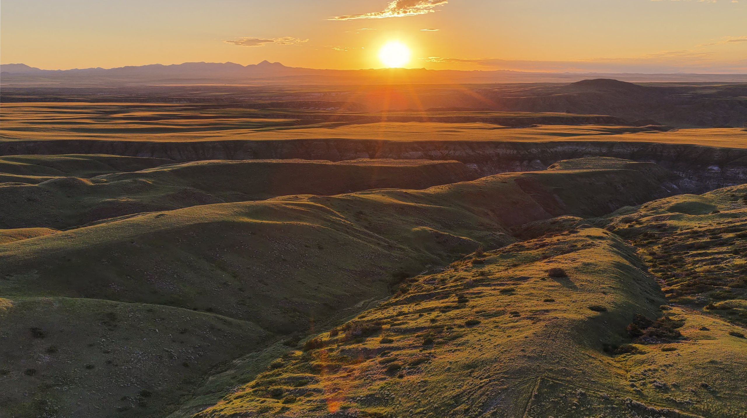 The sun sets over rolling green hills and distant mountains, casting long shadows and a golden glow across the landscape.