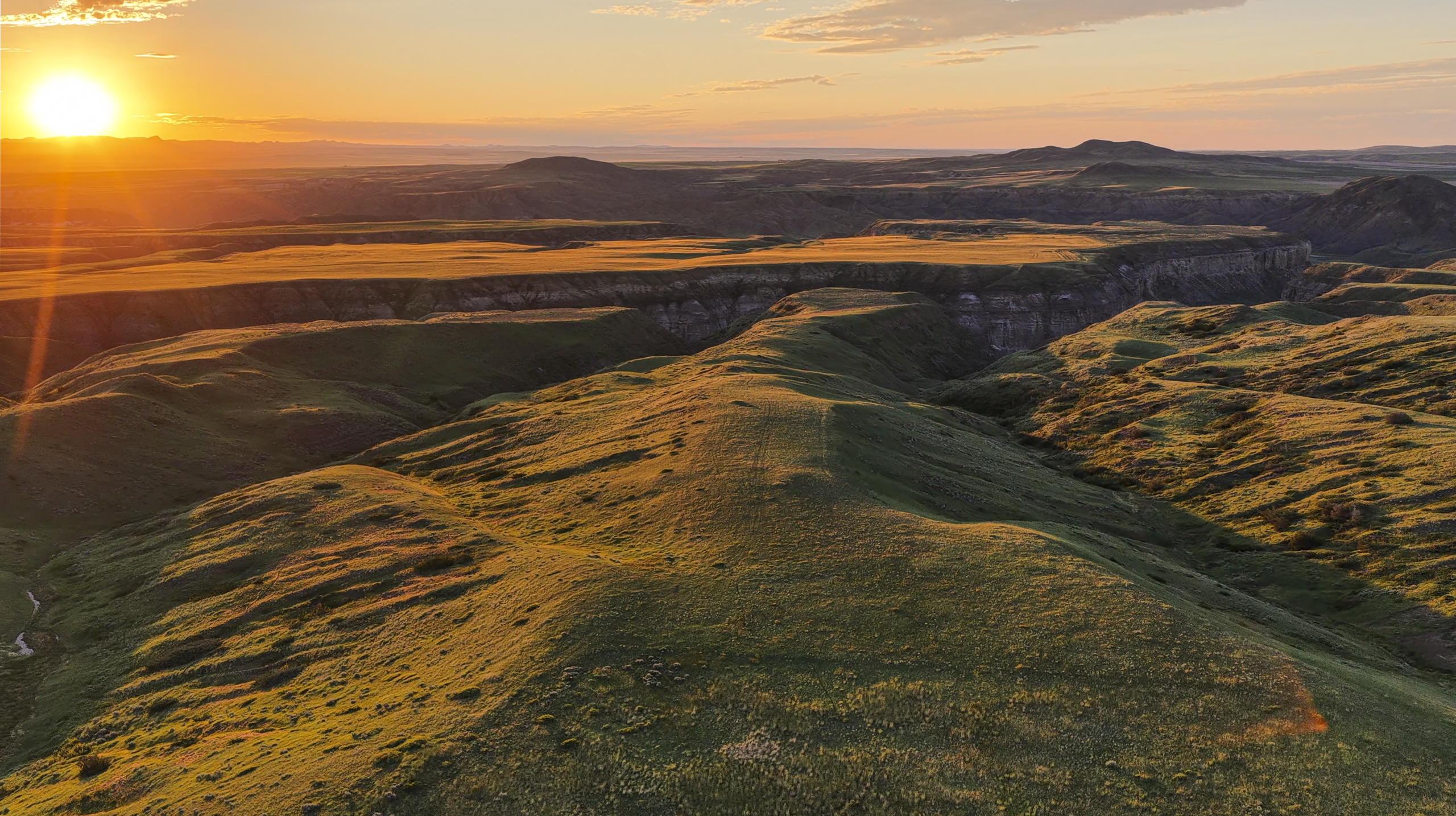 Sunset over grassy, rolling hills with deep valleys and distant mountains, casting long shadows and a golden glow across the landscape.
