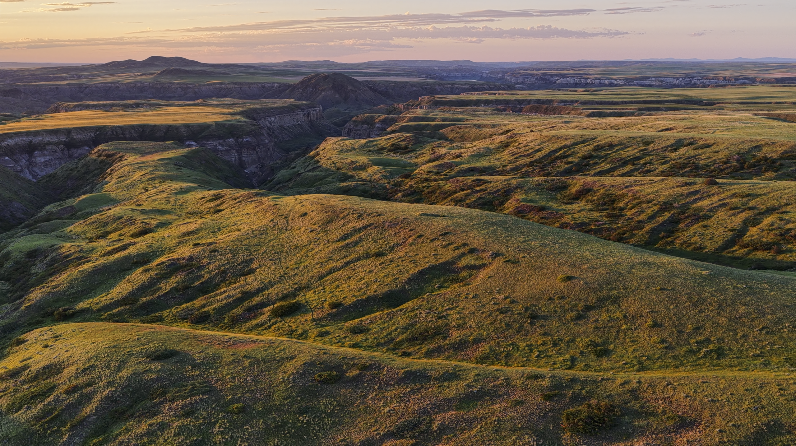 Aerial view of rolling green hills and deep valleys at sunset, with soft sunlight casting long shadows across the landscape. Distant plateaus and a clear sky are visible on the horizon.