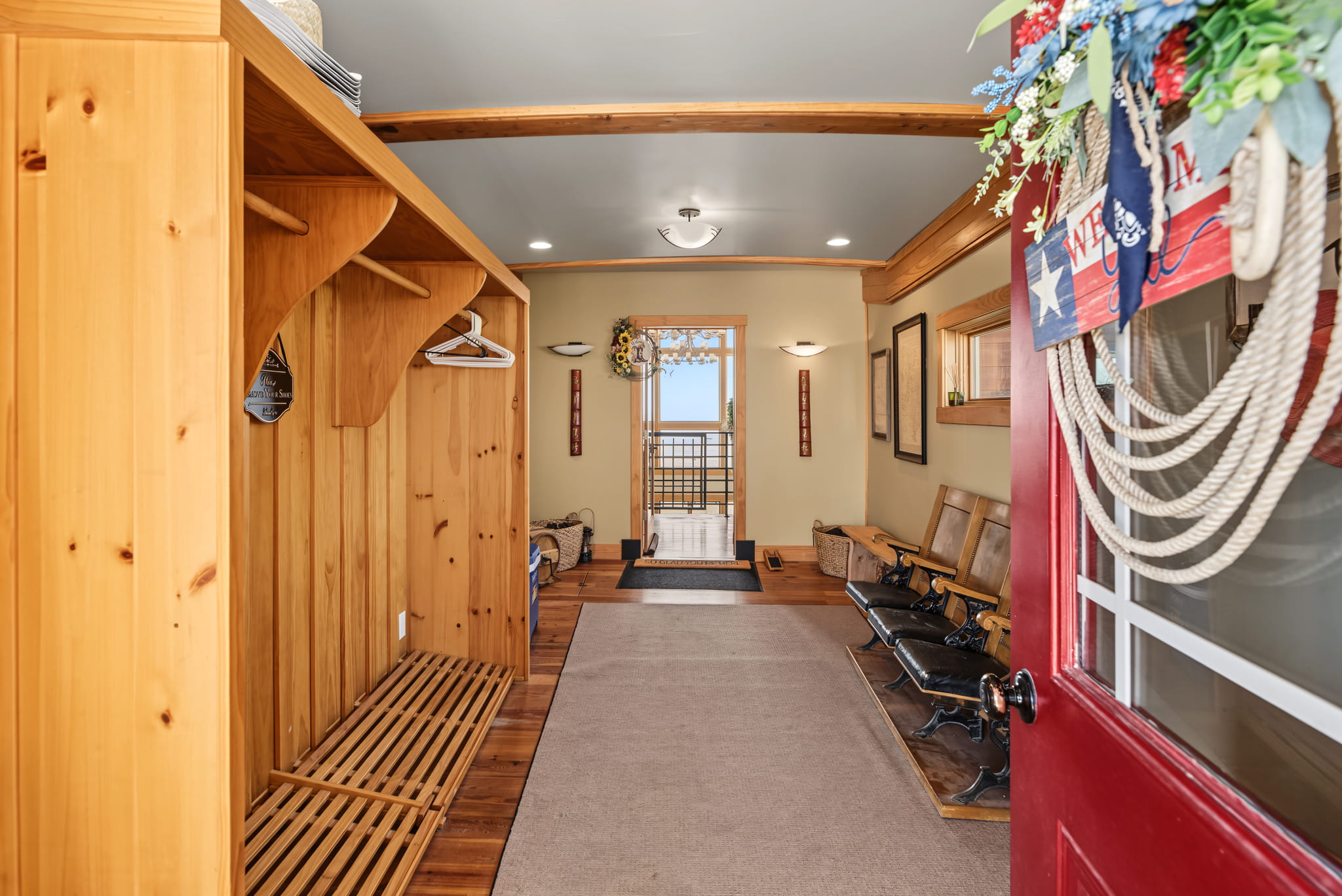 Red door opens to a hallway with wooden cubbies, benches, and a beige carpet. Decorations include flowers and a nautical rope wreath. A door at the end reveals an outside view.