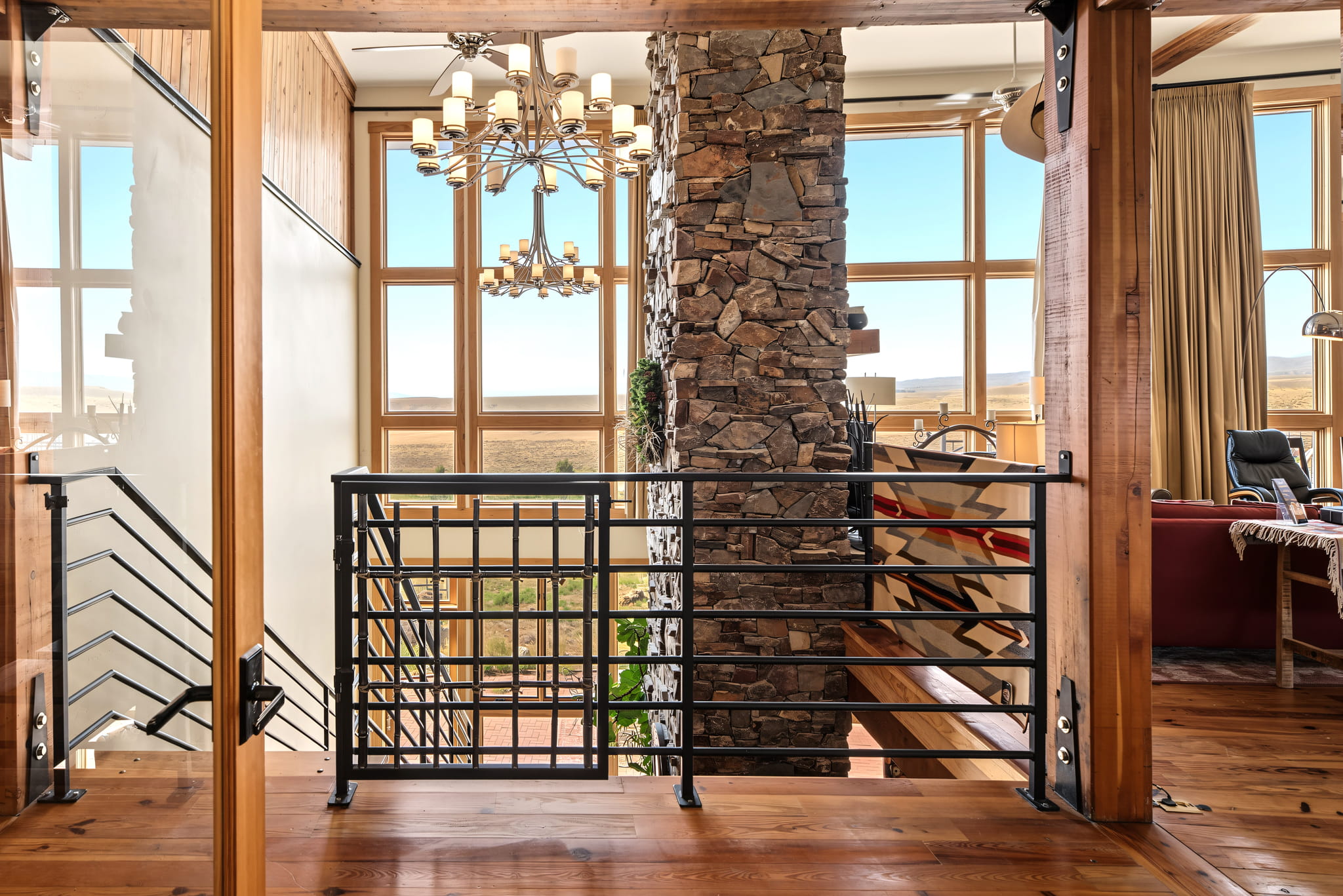 A cozy, modern interior with large windows, a stone column, wood floors, metal railing, and a chandelier. Natural light fills the space, and mountains are visible through the windows in the background.