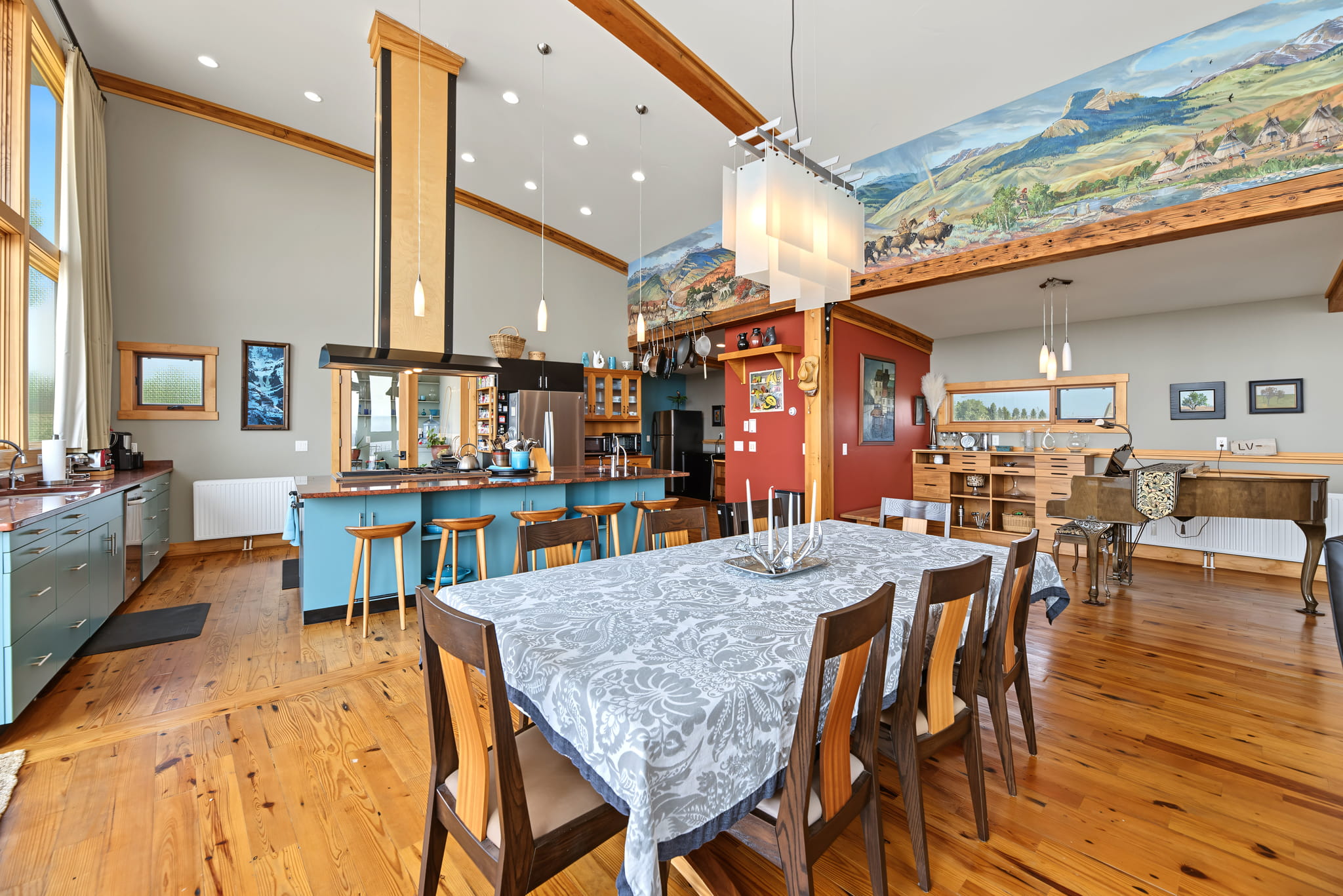 Open-concept kitchen and dining area with wooden floors, barstools at a blue island, a patterned tablecloth, large windows, and colorful mural along the upper wall. Natural light fills the spacious, modern room.