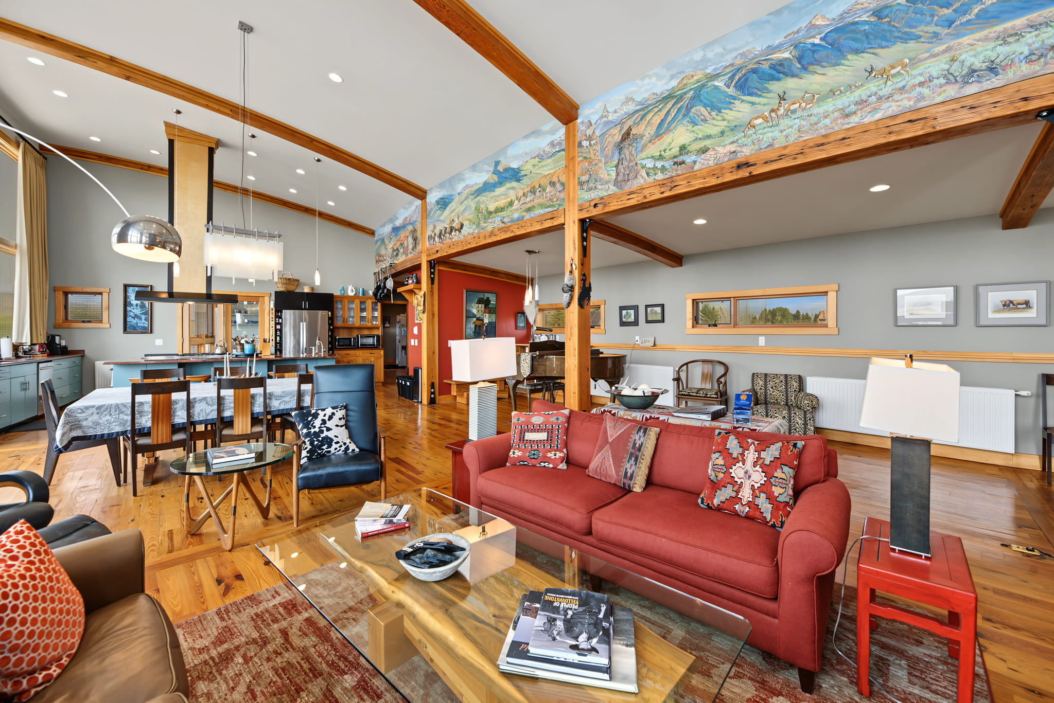 Spacious open-concept living area with a red sofa, dining table, modern kitchen, wood beams, hardwood floors, and a colorful mountain mural above the walls. Large windows bring in natural light.