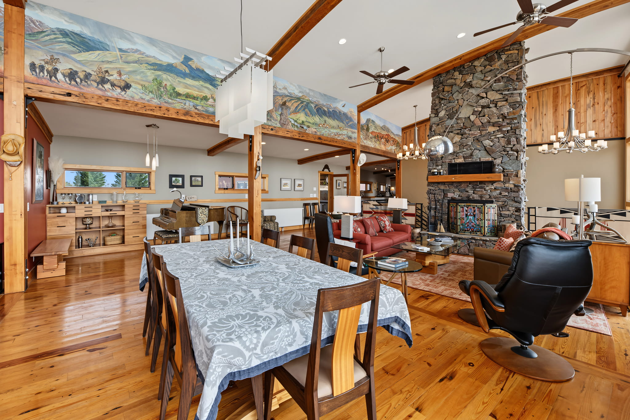 Open-concept living and dining area with wood floors, stone fireplace, murals above, large windows, modern furniture, and rustic decor. Dining table with a gray tablecloth is in the foreground.