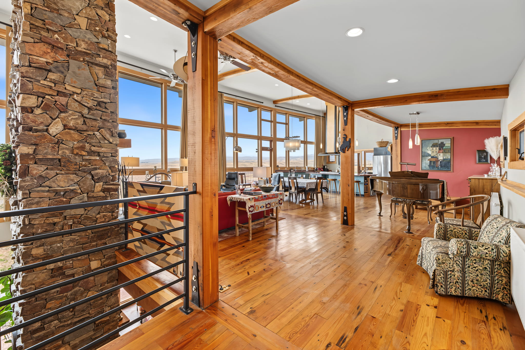 Spacious, modern living and dining area with large windows, wooden floors, exposed beams, stone pillar, piano, patterned armchairs, and red accent wall, overlooking a scenic outdoor view.