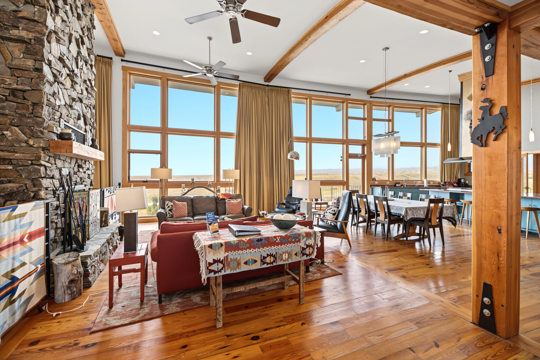 Spacious, sunlit living and dining area with tall windows, stone fireplace, wooden floors, and ceiling beams. Southwestern decor, comfortable seating, and a modern kitchen are visible.