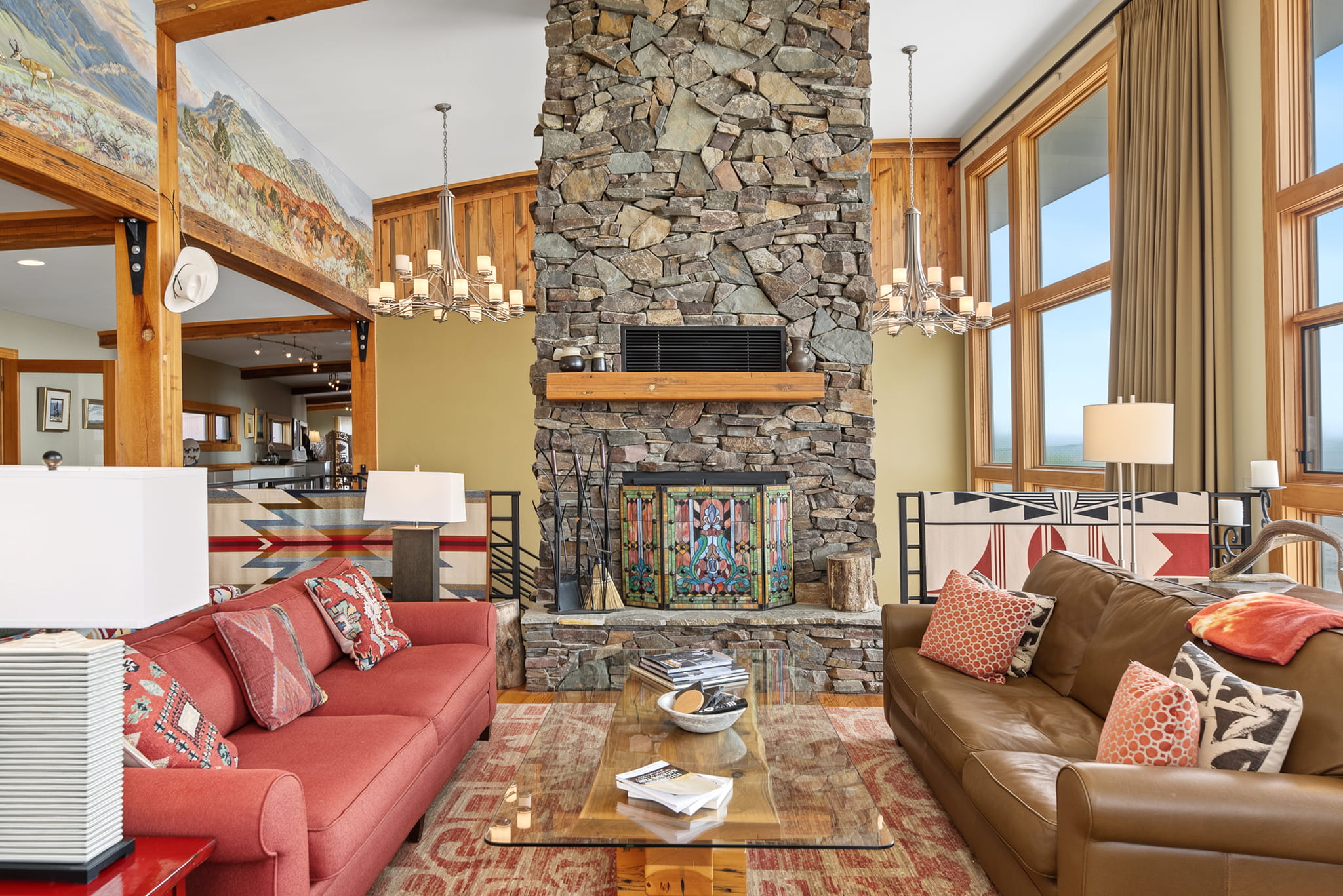 A cozy living room features a stone fireplace, large windows, a brown leather couch, a red sofa, colorful pillows, a glass coffee table, chandeliers, and wooden beams with mural artwork above.