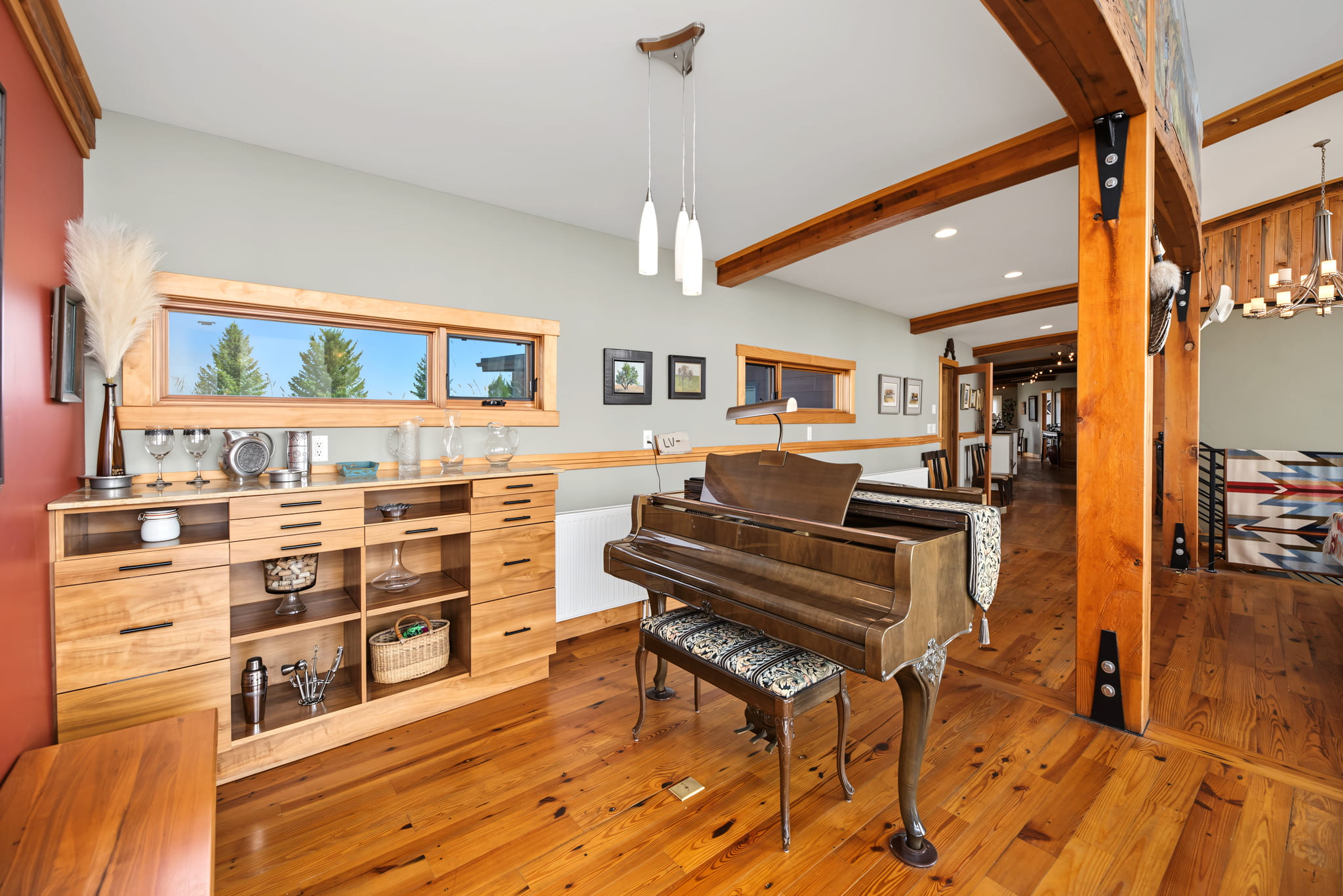 A bright room with a wooden floor, a grand piano with a patterned bench, wooden shelves, pendant lights, and large windows showing trees outside. A long hallway with artwork is visible in the background.