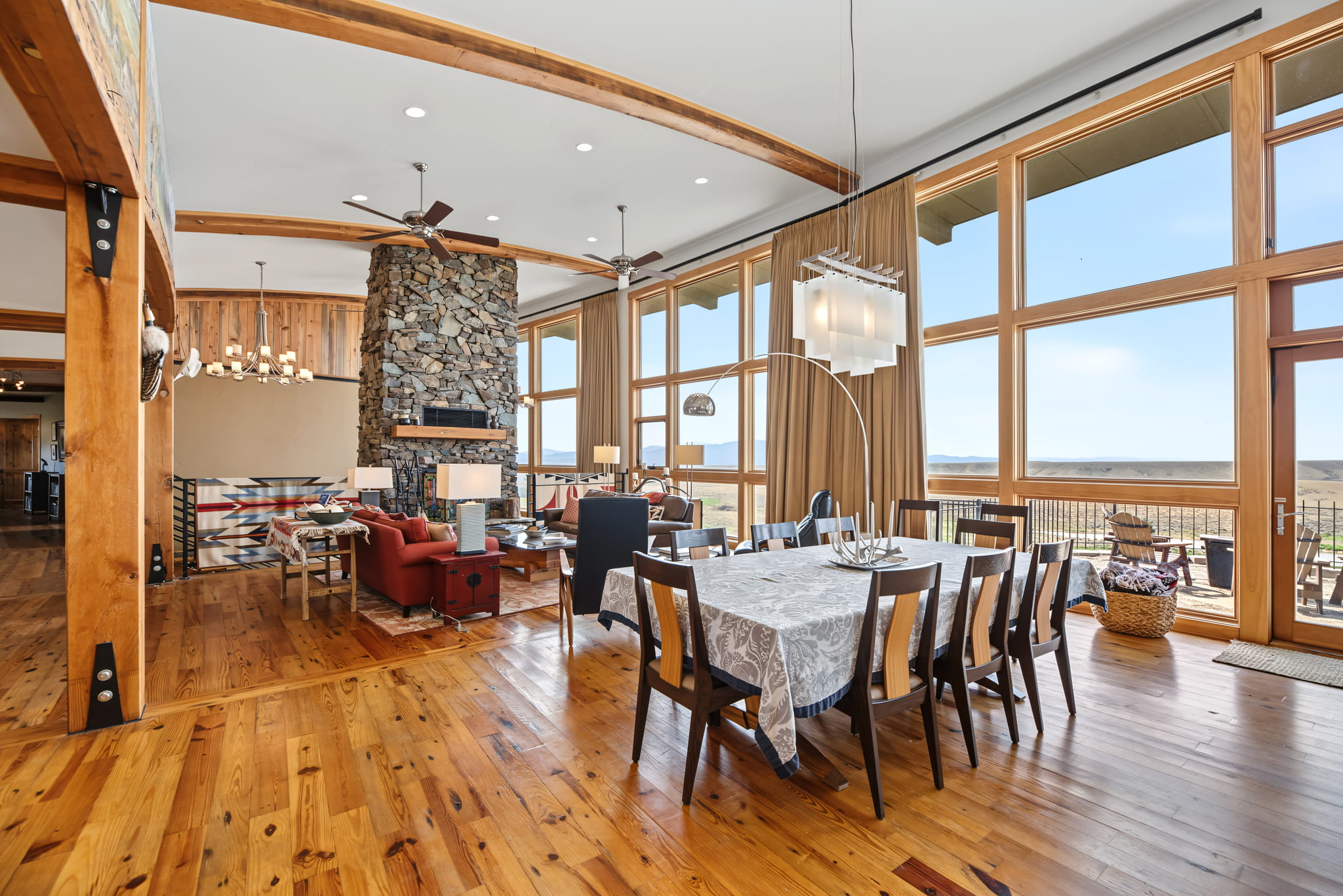 Spacious open-concept living and dining area with wood floors, large windows, rustic beams, stone fireplace, and modern furnishings. Dining table seats eight under a contemporary light fixture.