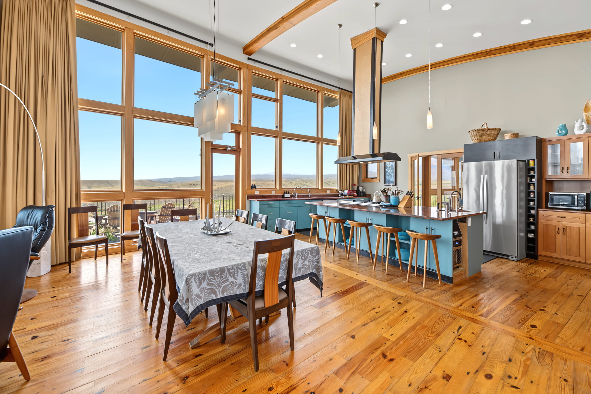 Spacious modern kitchen and dining area with large windows, wooden floors, a long table with a gray tablecloth, blue island with stools, and stainless steel appliances.