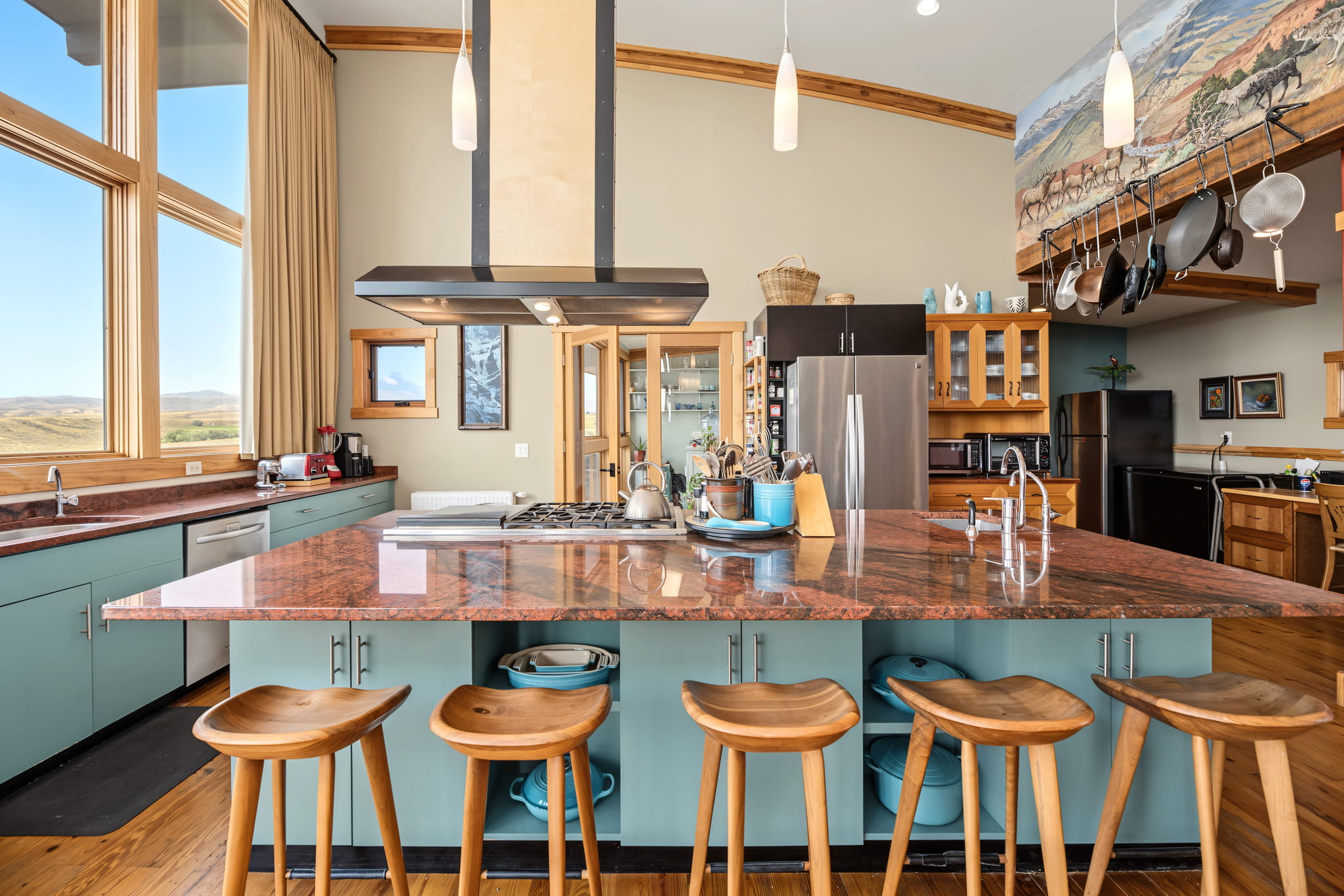 Spacious kitchen with turquoise cabinets, red granite island, wooden stools, stainless steel appliances, large windows, and a mural above a wood-paneled area. Natural light highlights modern and rustic design elements.