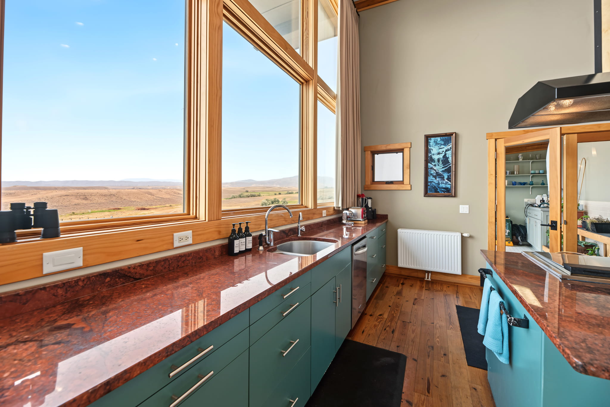 Modern kitchen with teal cabinets, red granite countertops, wooden floors, large windows offering a view of rolling hills and blue sky, and various kitchen essentials neatly arranged.