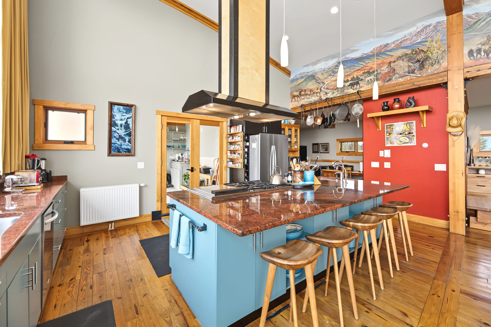 Spacious kitchen with a large island, wooden stools, hanging pots, red accent wall, light wood floors, colorful mural above cabinets, modern appliances, and ample natural light.
