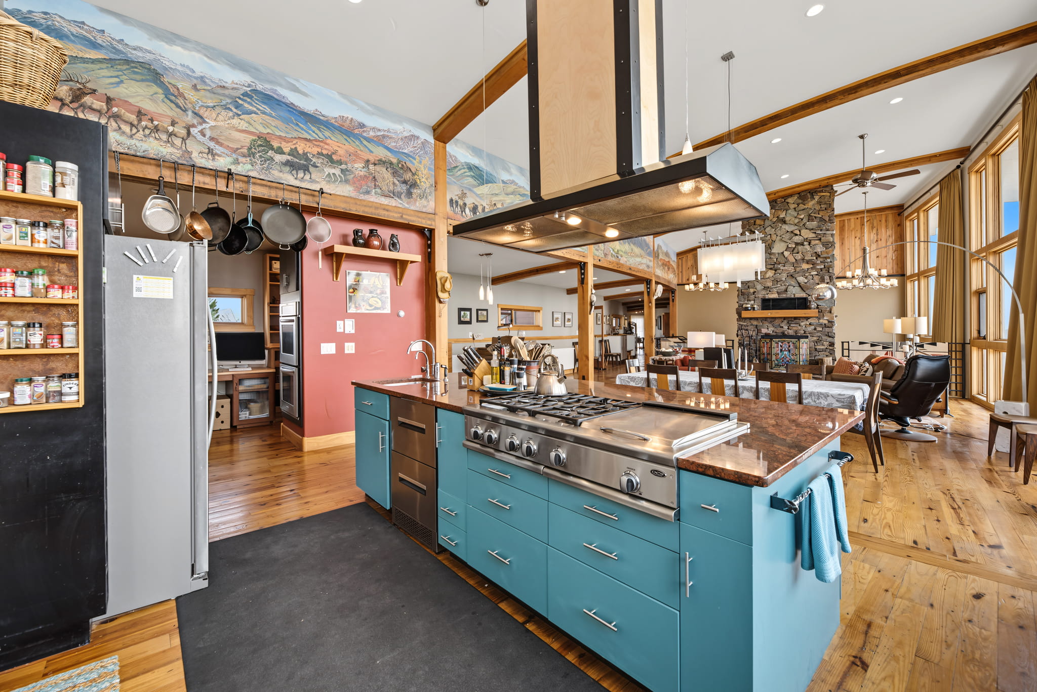 Spacious kitchen with a blue island, gas stove, hanging pots, stainless steel fridge, wood floors, large windows, stone fireplace, and colorful mural above. Open layout connects kitchen, dining, and living areas.