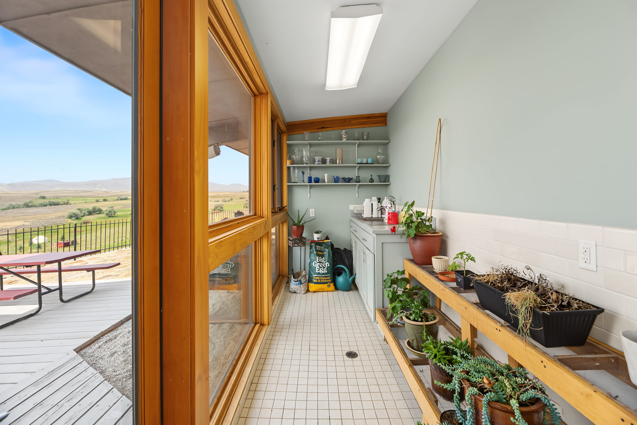 A sunlit room with large windows, plants on a wooden bench, cabinets with dishes, and a view of a deck and open fields outside.