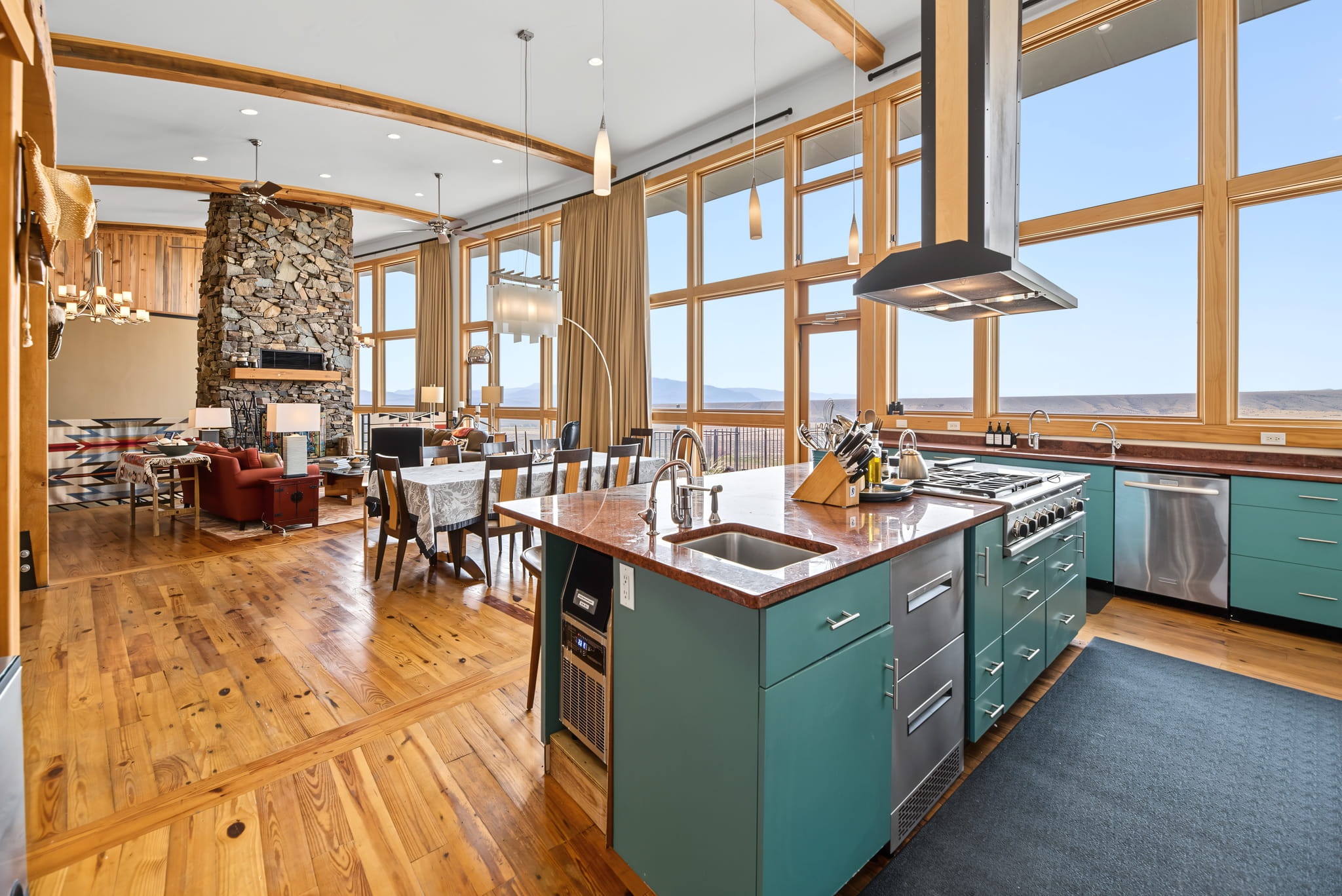 Modern open-concept kitchen with teal cabinets, wood floors, stainless steel appliances, large windows, dining area, and a living room with a stone fireplace. Mountain views can be seen through the windows.