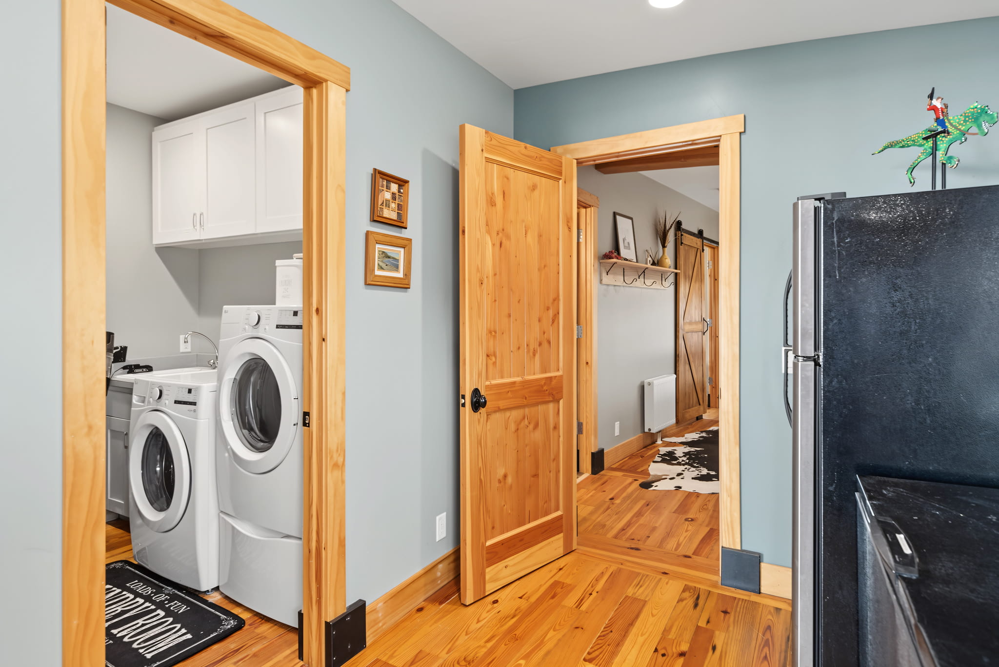 A laundry room with a front-load washer and dryer, wooden doors, white cabinets, and light blue walls. A black refrigerator is partially visible, and a hallway with wooden floors and a coat rack is seen.