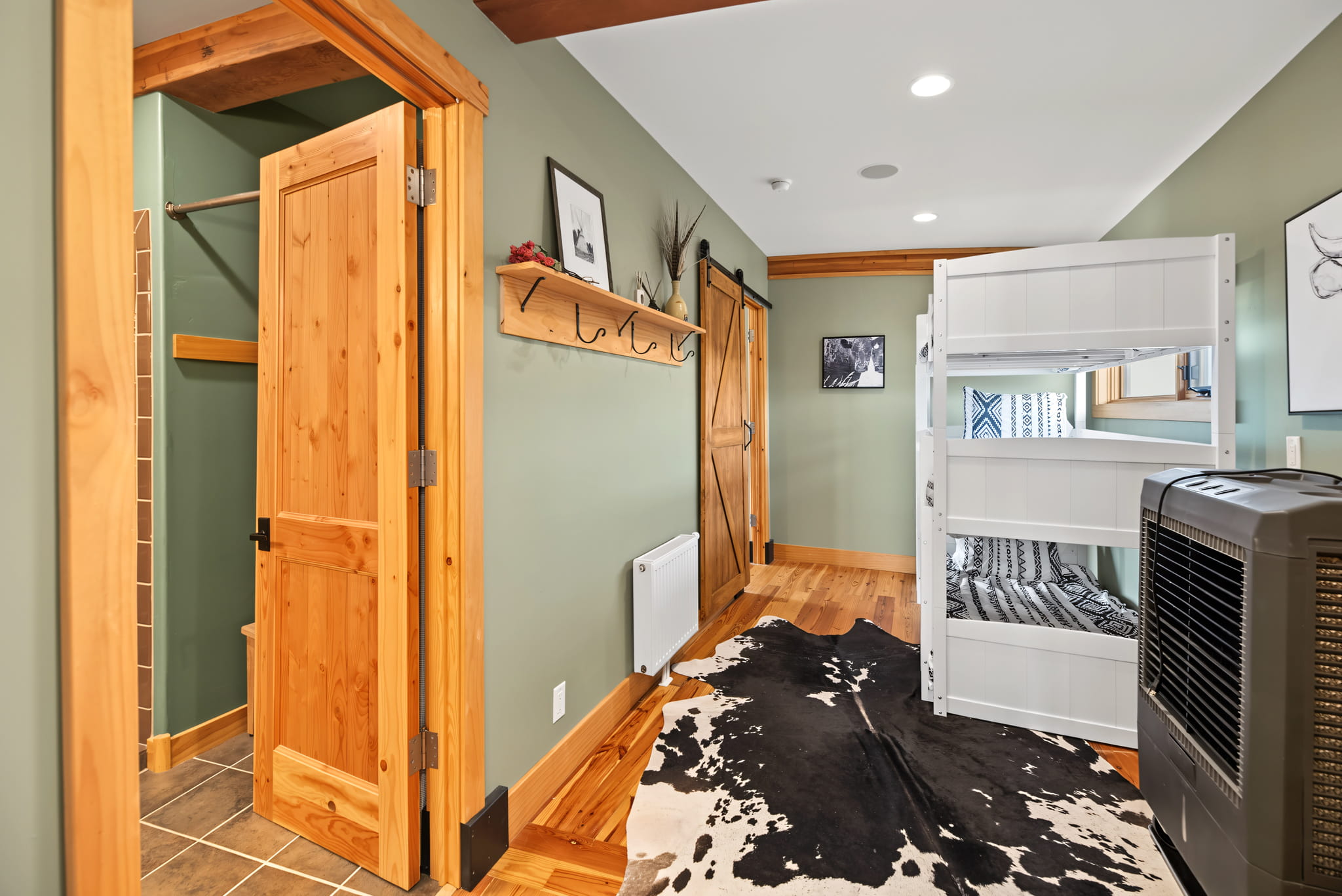 A cozy room with green walls, wooden doors, a sliding barn door, white bunk beds, a cowhide rug, coat hooks, framed art, and a small heater on a hardwood floor.