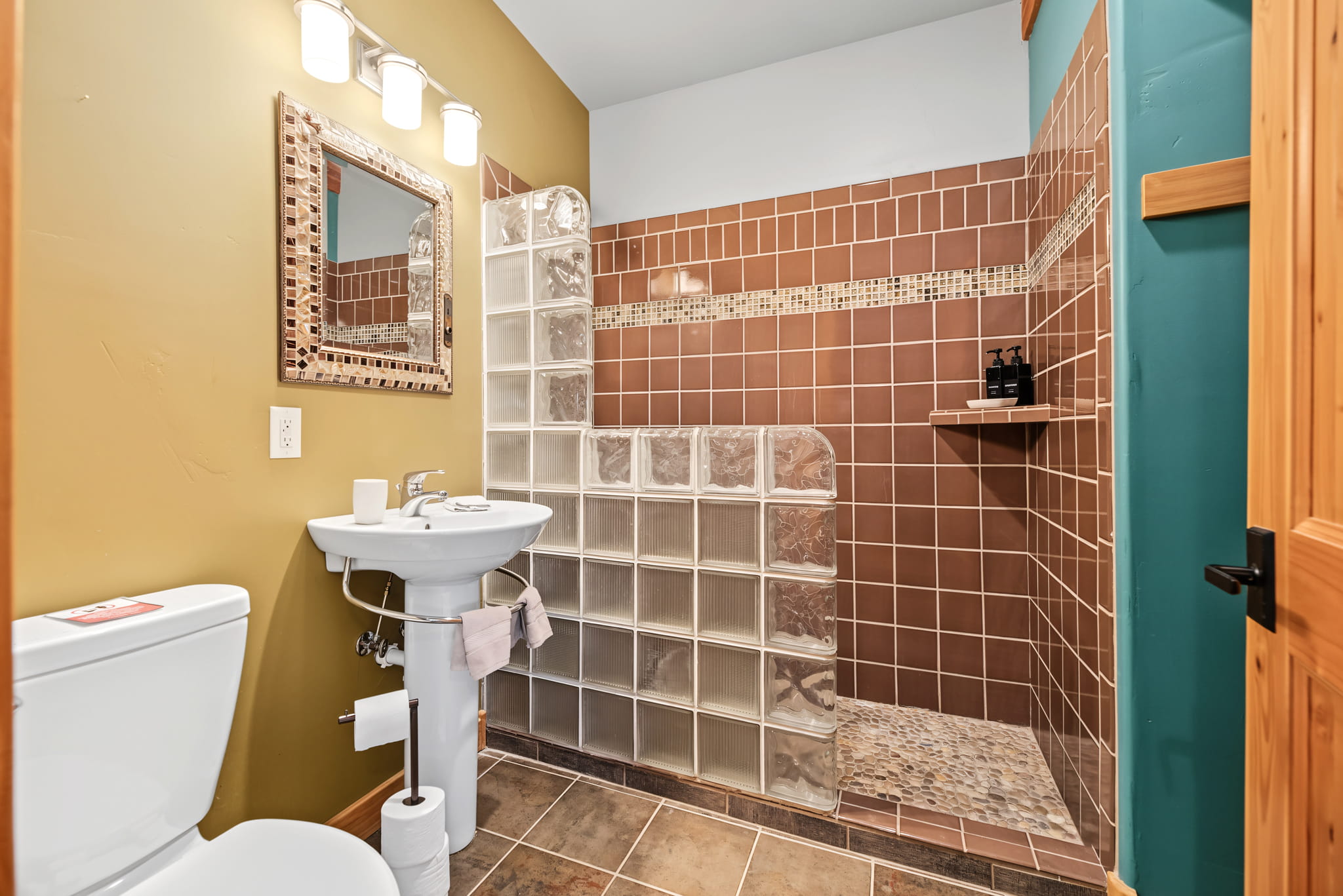 Bathroom with pedestal sink, mirror, toilet, and open glass-block walk-in shower with brown tiles and mosaic accent. Wall is painted yellow and teal; wood door is partially visible.