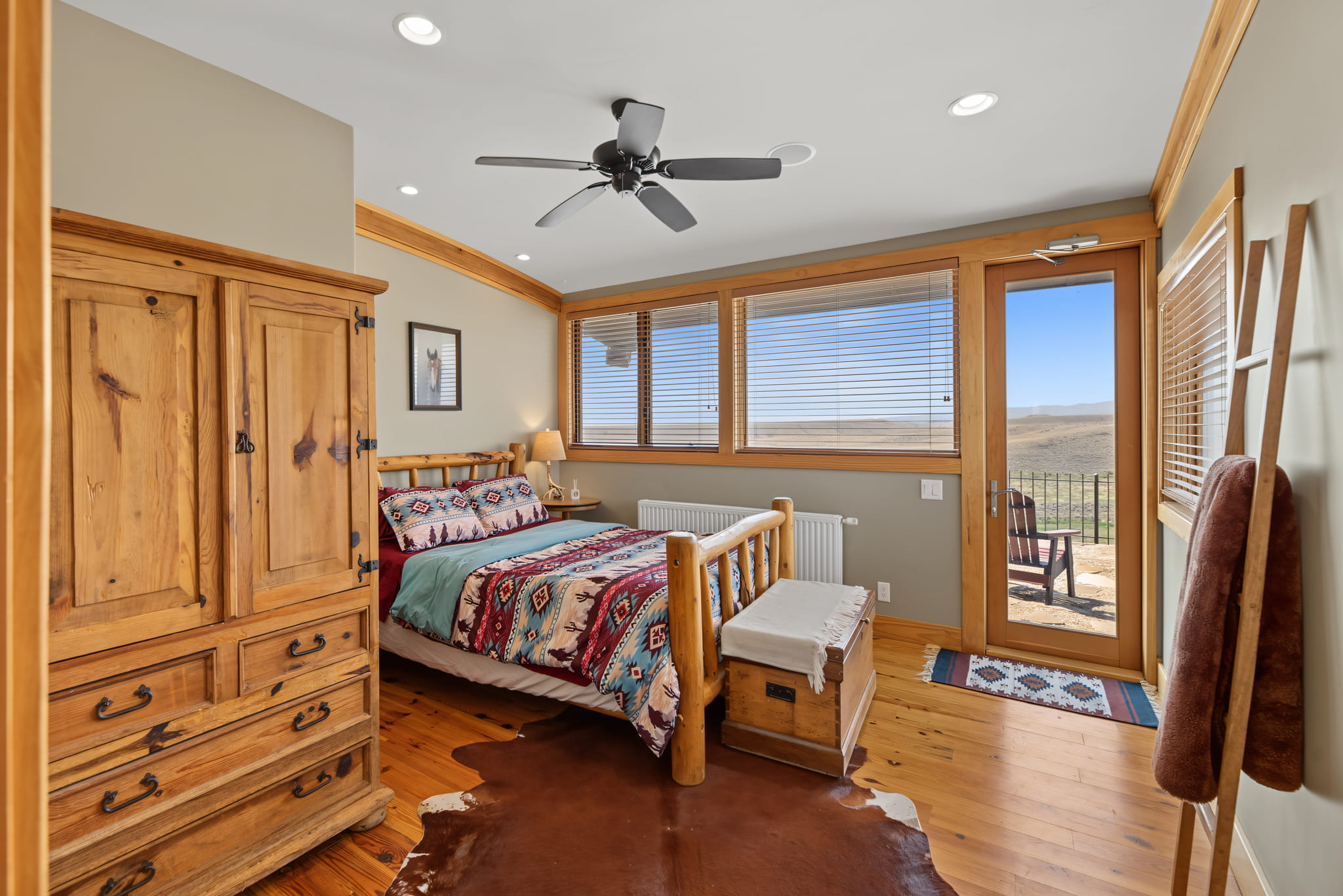 Cozy bedroom with wood furniture, patterned bedding, and large windows offering a scenic outdoor view. A ceiling fan, cowhide rug, and a wooden chest add rustic charm to the space.