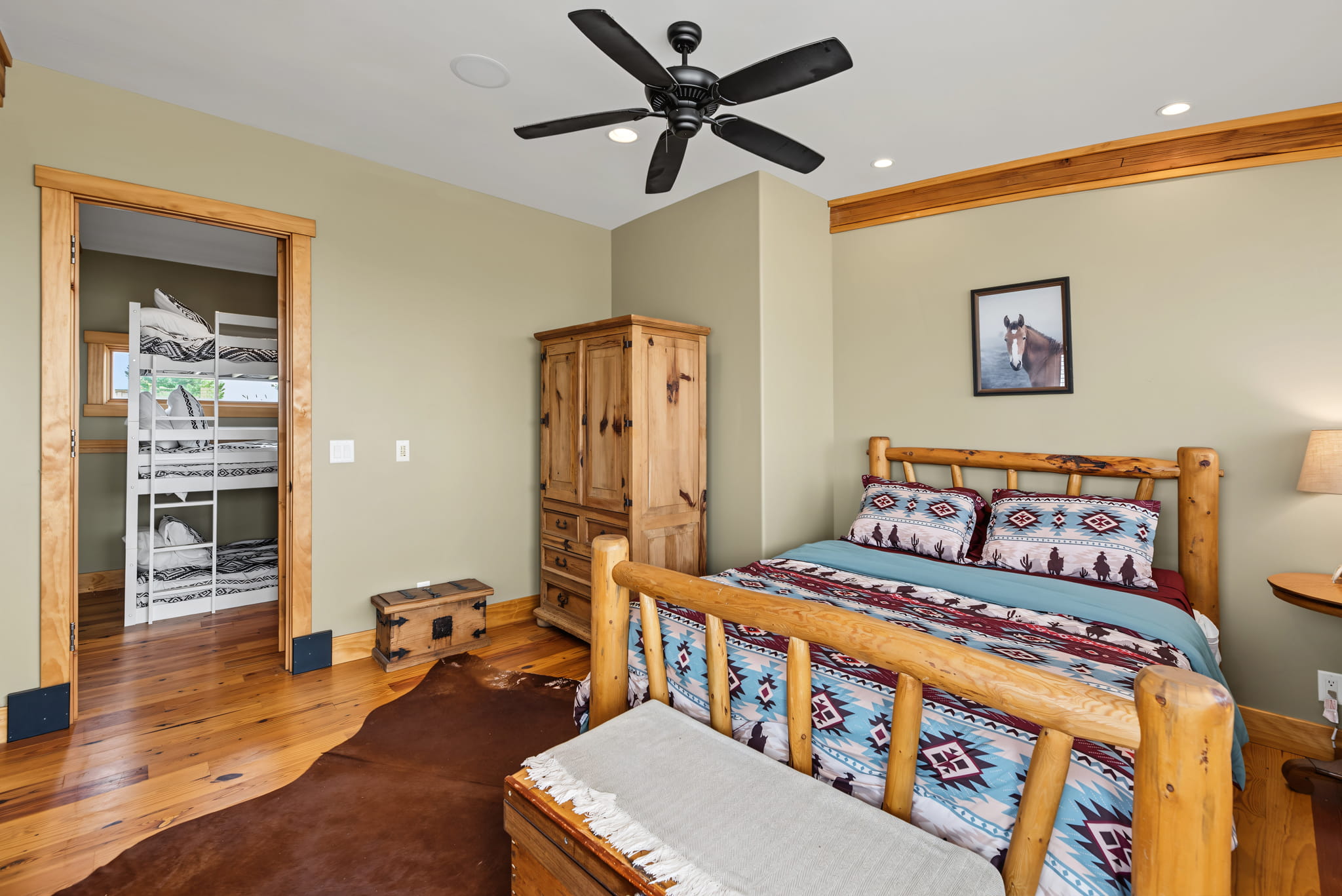 Rustic bedroom with log furniture, patterned bedding, wooden floors, a cowhide rug, ceiling fan, horse photo on wall, and an open door showing white bunk beds in the next room.