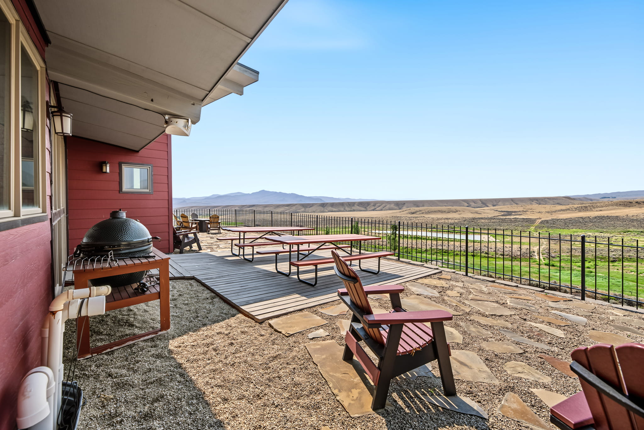 Red house patio with Adirondack chairs, picnic tables, and a grill, overlooking a wide, open landscape with hills under a clear blue sky. Iron fence separates patio from the view.