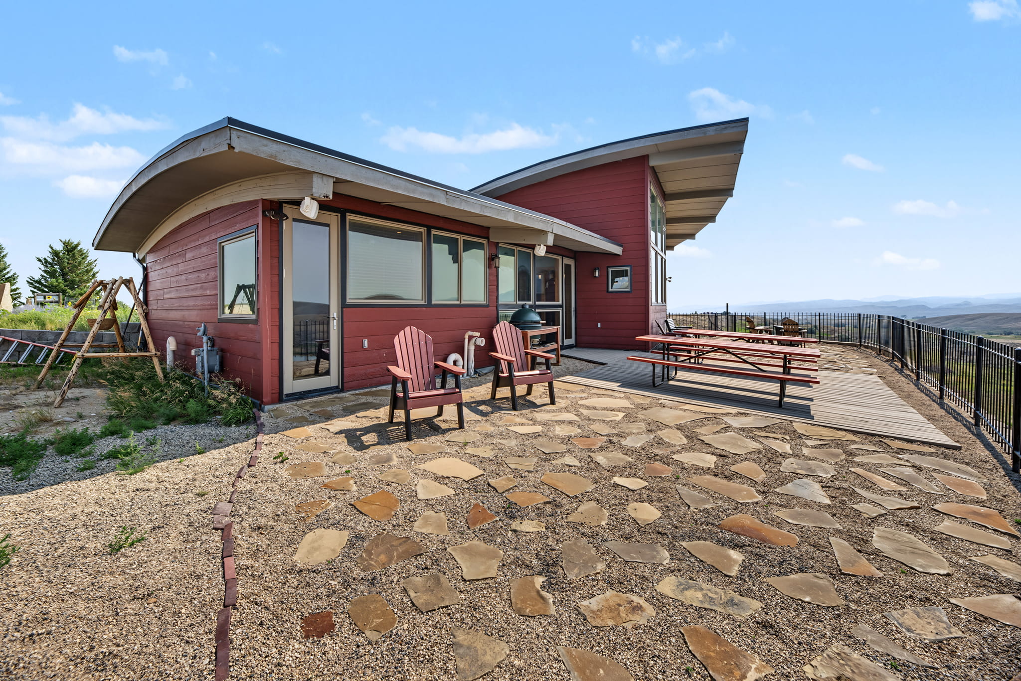Modern red house with curved roof, large windows, and a wooden deck overlooking a scenic landscape. Two Adirondack chairs and a picnic table are on a stone patio in the foreground under a clear, blue sky.