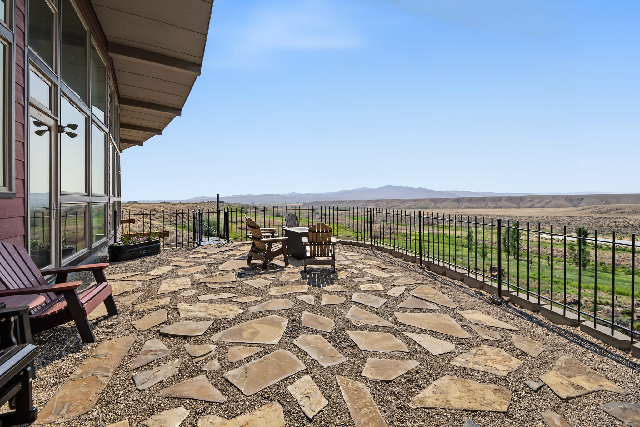 Stone patio with Adirondack chairs overlooks a scenic desert landscape and distant mountains, bordered by a metal fence and next to a modern house with large windows under a clear blue sky.