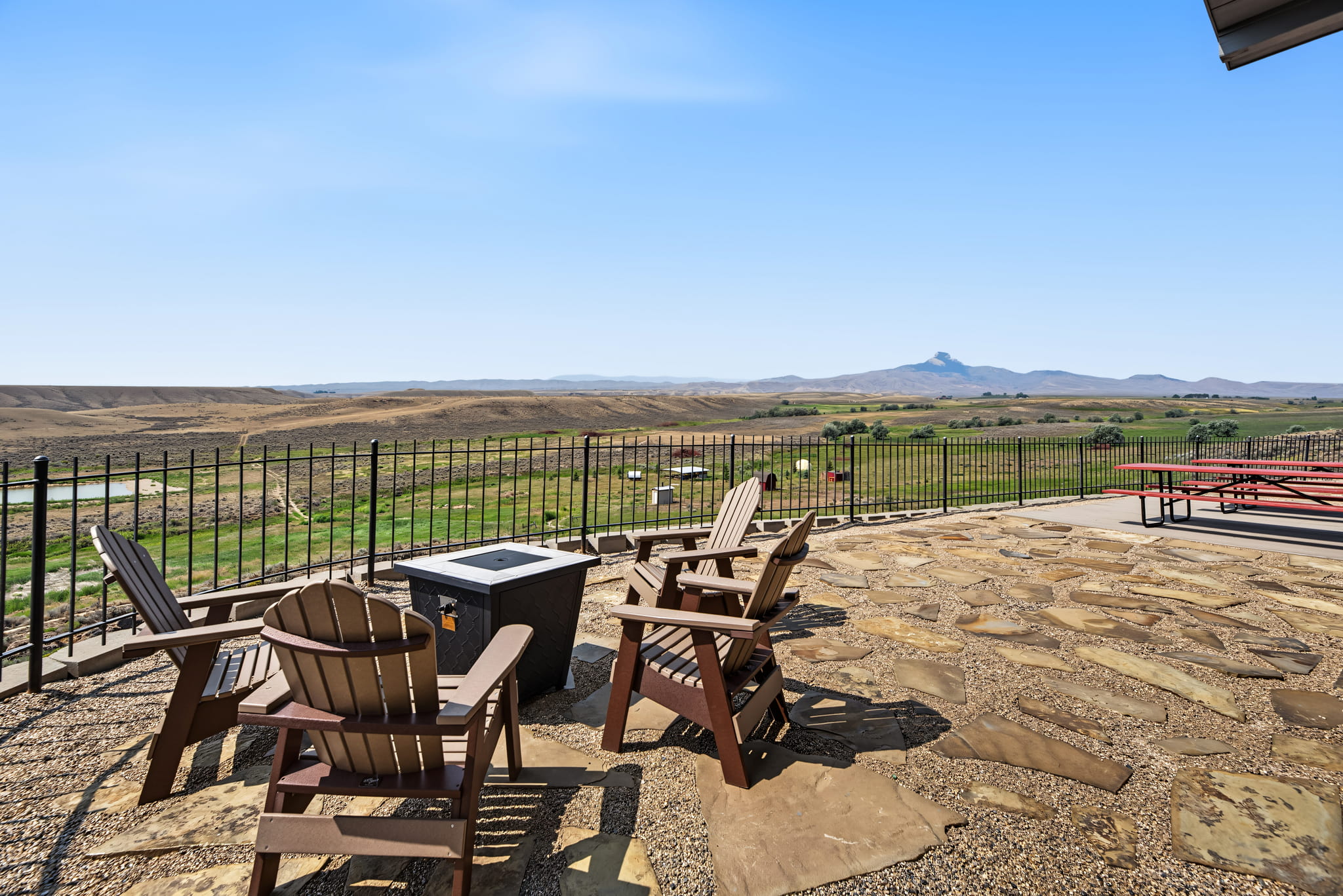 Four wooden chairs surround a small fire pit on a stone patio overlooking a scenic, open landscape with hills, green fields, and distant mountains under a clear blue sky.