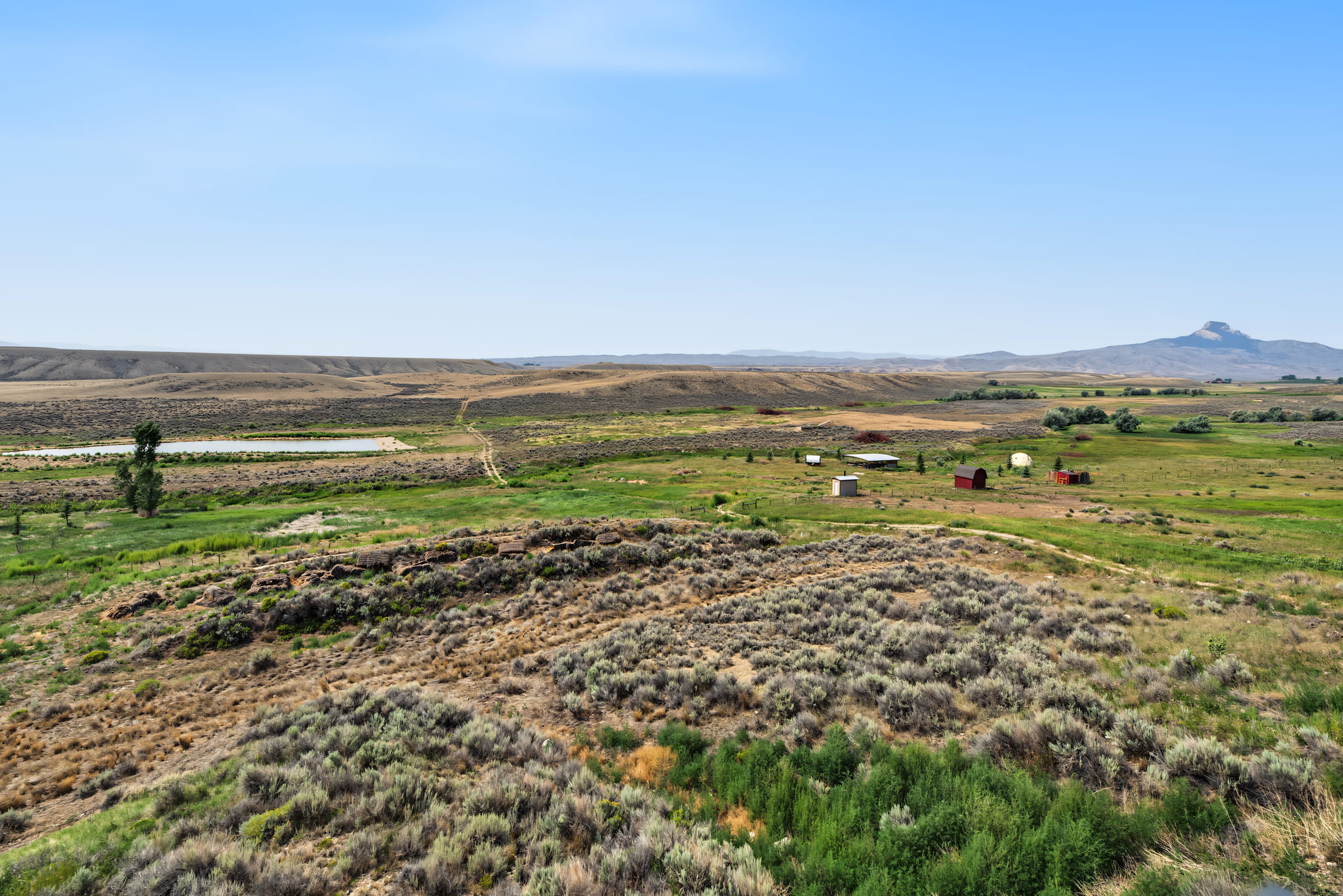 Wide view of a rural landscape with grassy fields, scattered shrubs, small buildings, and distant low mountains under a clear blue sky.
