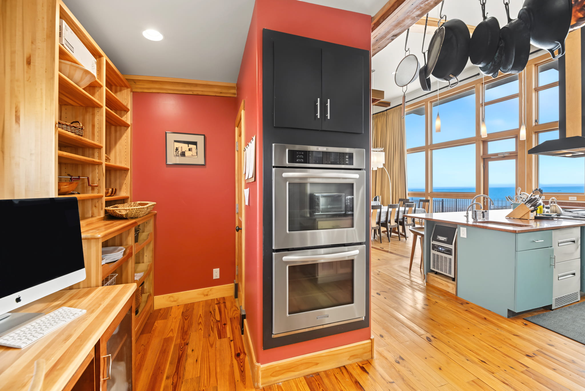 Modern kitchen with double wall ovens, wood floors, built-in shelves, a computer workstation, and large windows offering a scenic ocean view. Pots hang above a blue island in the center of the space.