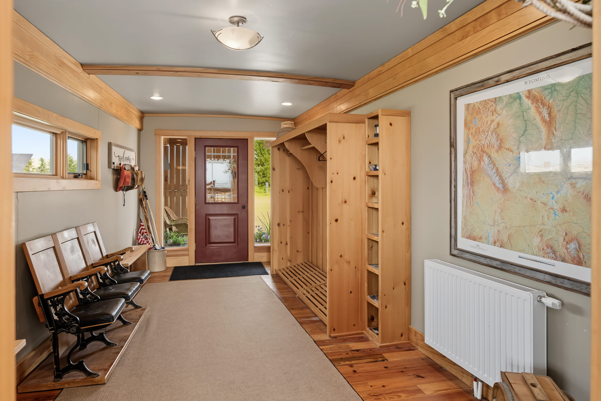 Wooden entryway with a bench, coat hooks, storage cubbies, and a large wall map. Light streams in from windows; a maroon door leads outside. Ceiling lights and light wood trim create a warm, inviting atmosphere.